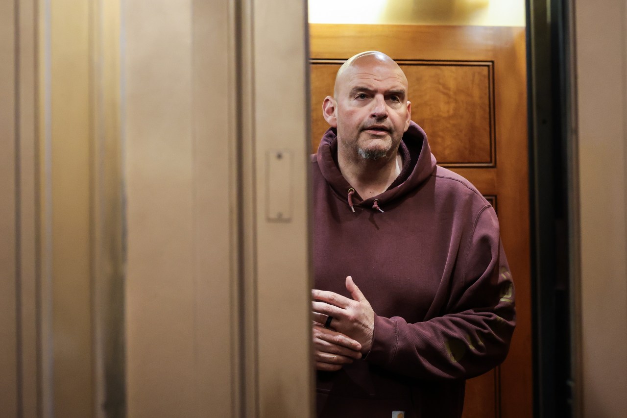 Bald man with goatee in a maroon hoodie standing in an elevator doorway inside the U.S. Capitol, hands clasped in front of him.