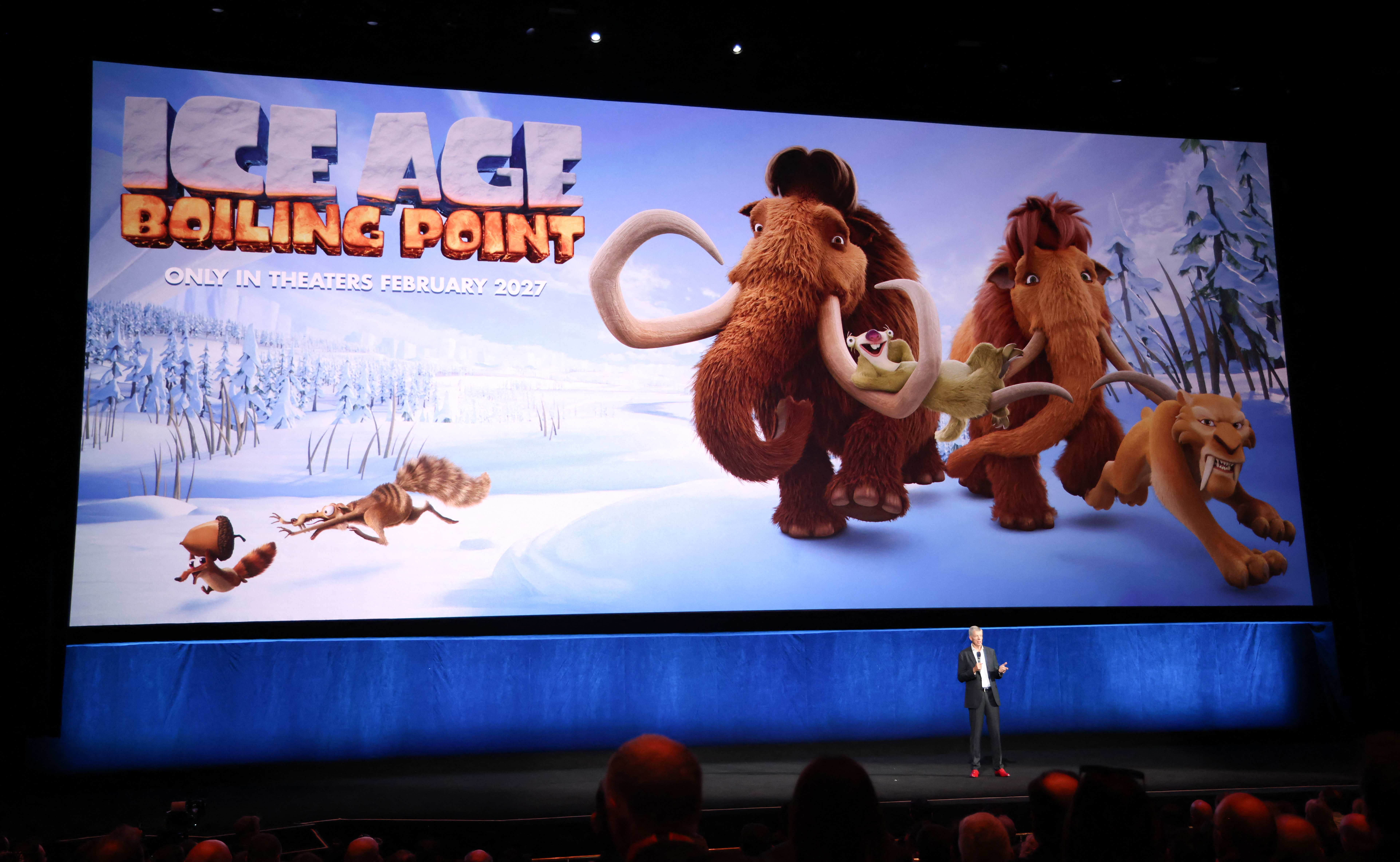A presenter stands on a darkened stage beneath a giant 'Ice Age: Boiling Point' movie poster showing animated mammoths, a saber-tooth tiger, and a squirrel at CinemaCon.
