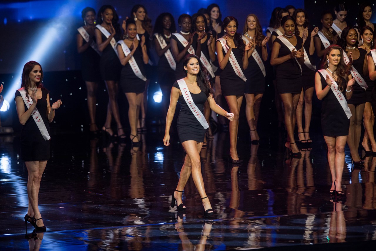 Woman in a black dress and 'United States' sash walks a reflective stage surrounded by other pageant contestants at Miss World 2016.
