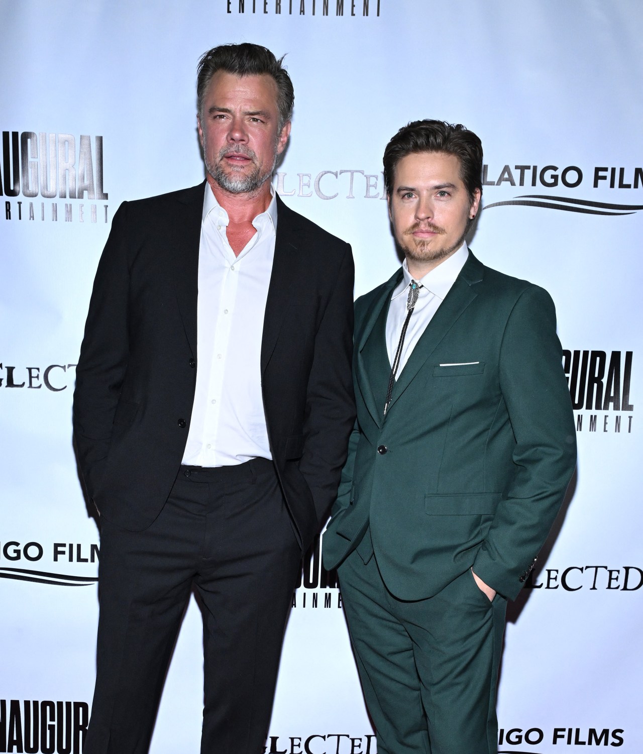 Two men in suits posing on a promotional backdrop — one in black and white, the other in a teal-green suit with a bolo tie.