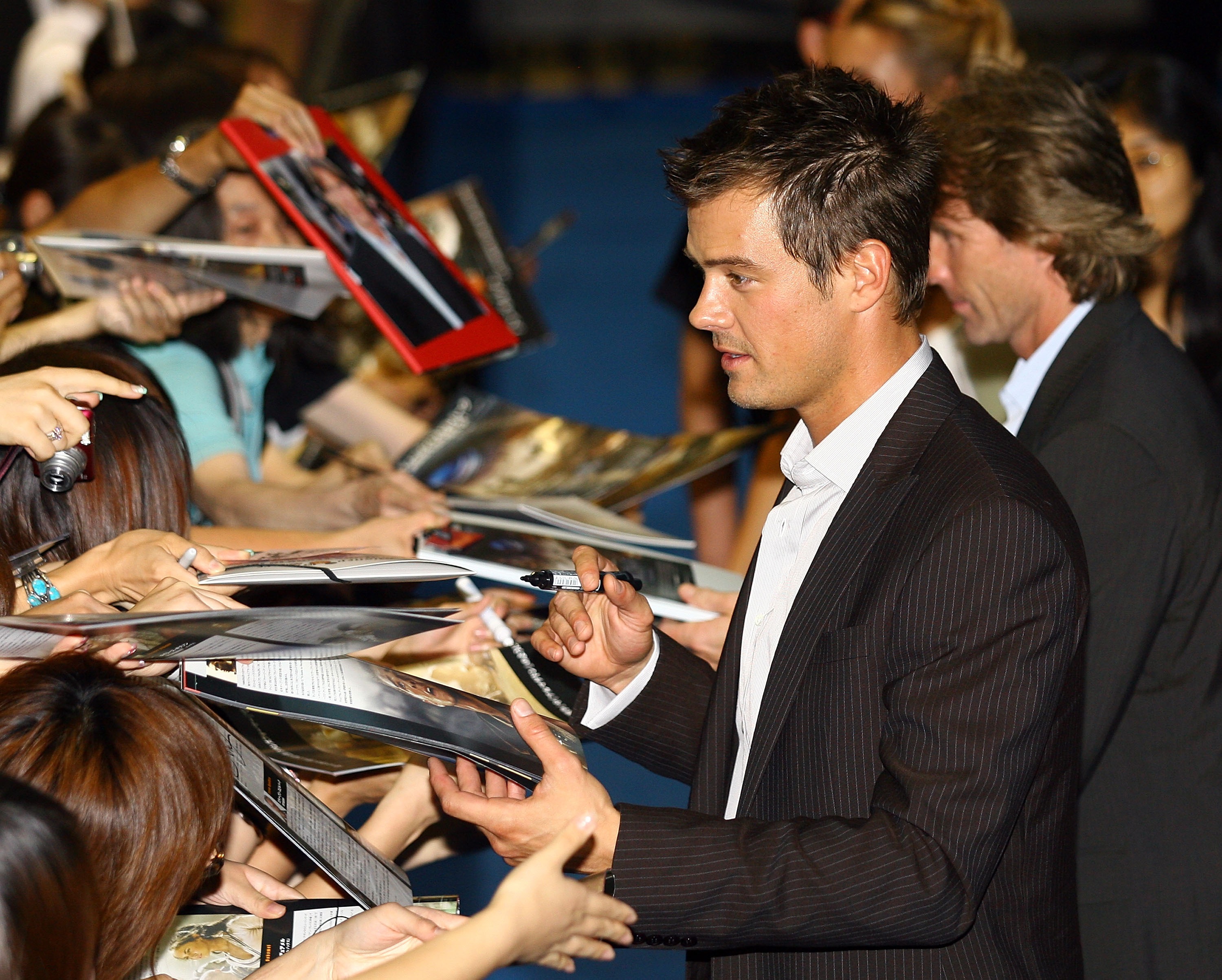 Click to open the image gallery A man in a dark pinstripe blazer signing autographs for a large crowd of fans at a movie premiere in Tokyo.