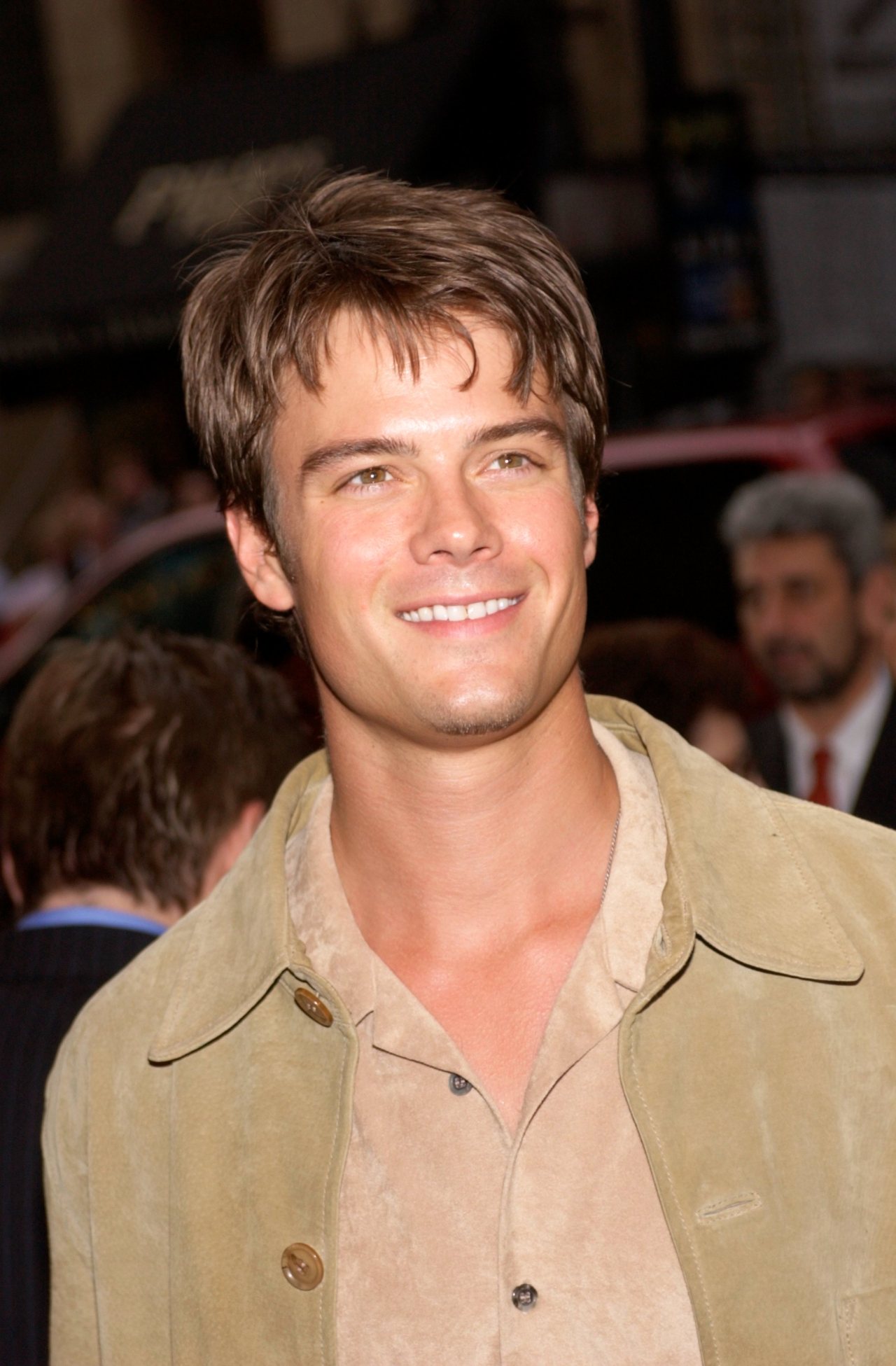 Josh Duhamel smiling at a public event, wearing a tan suede jacket over a beige shirt, with a crowd blurred in the background.