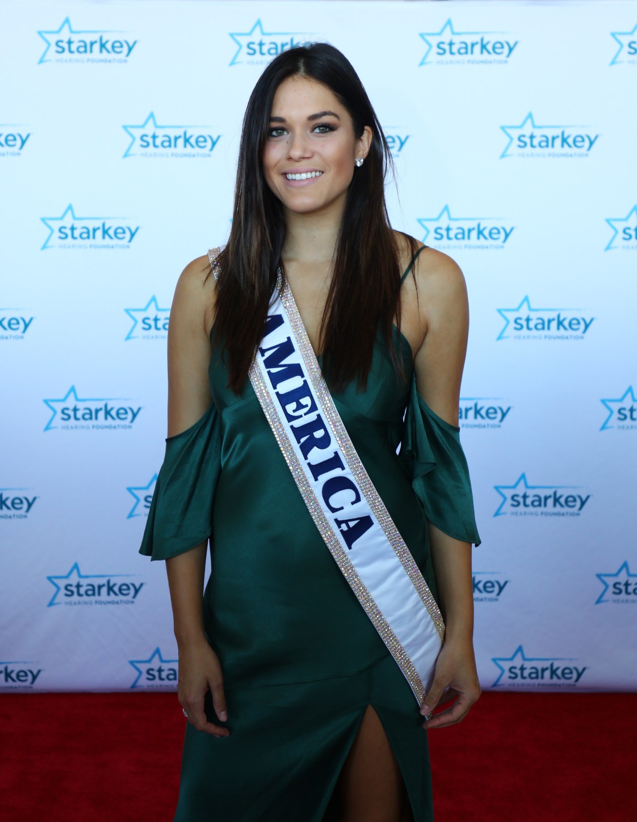 Young woman in a green gown with a rhinestone-trimmed 'America' sash smiling at the Starkey Hearing Foundation gala red carpet.