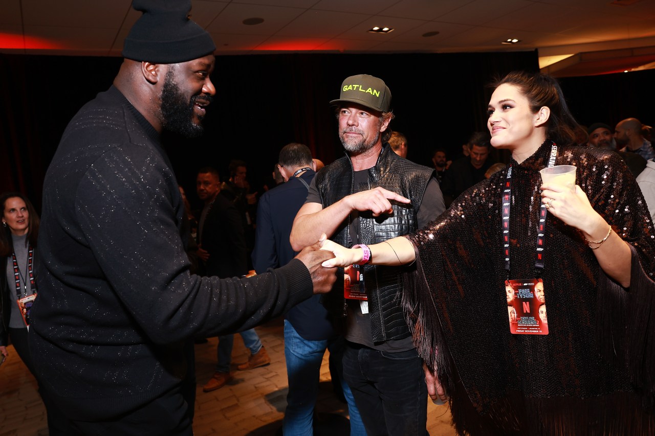 Shaquille O'Neal shakes hands with a woman while a man in a cap and leather vest looks on at an indoor event venue.