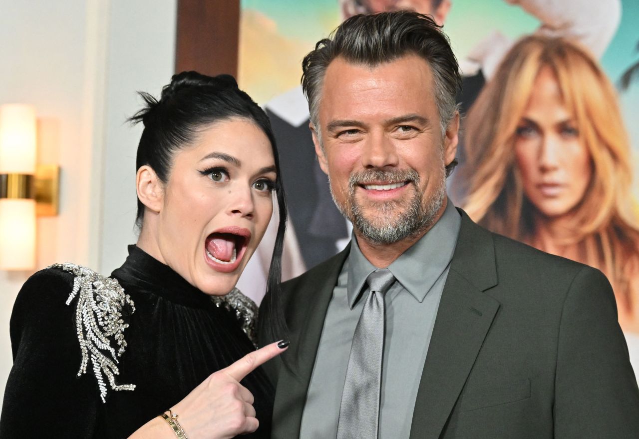 Woman in a black embellished top making a surprised face and pointing at a smiling man in a grey suit at a movie premiere.