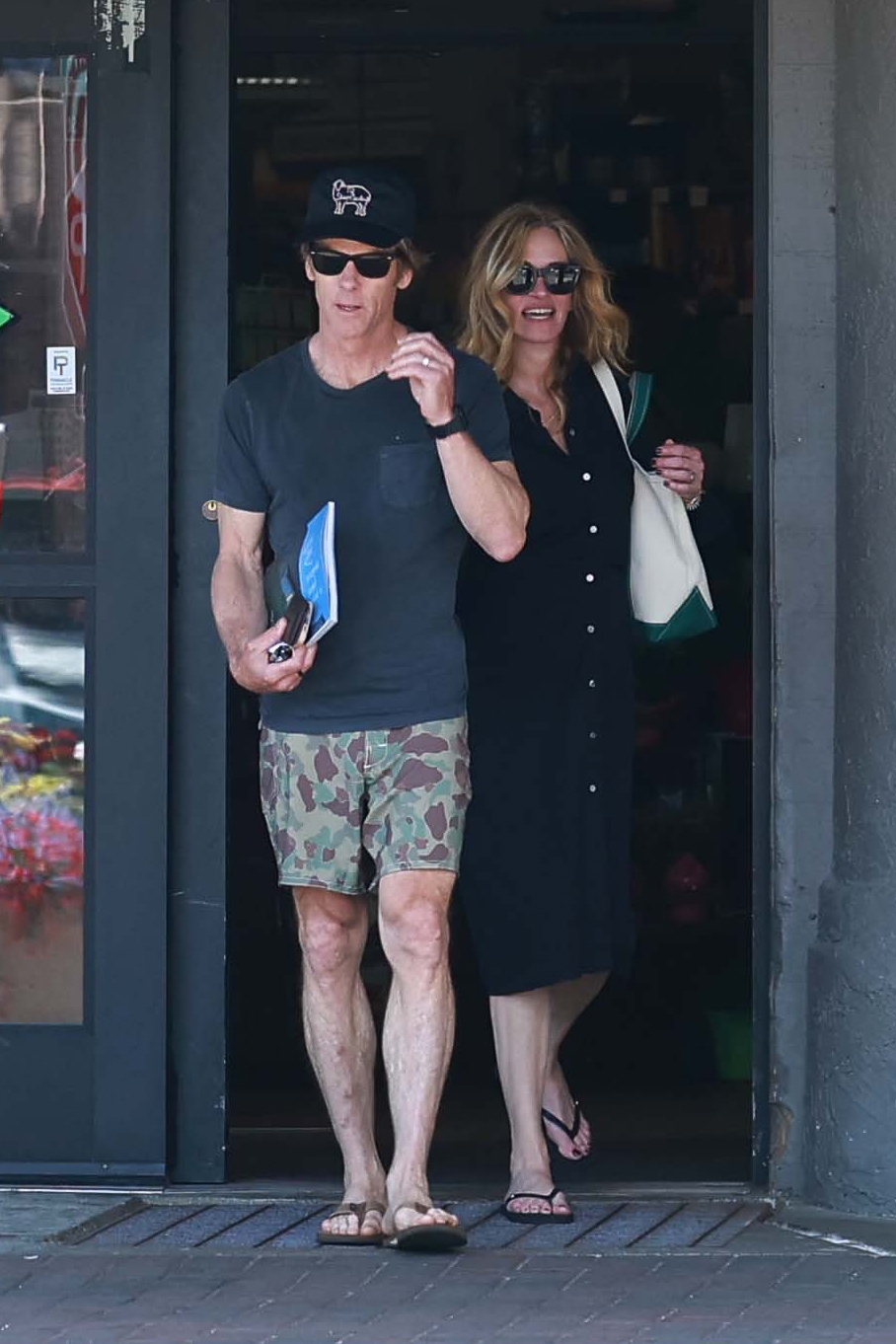 A man in a black cap, navy t-shirt, and camo shorts walks alongside a blonde woman in a black button-down dress, both wearing sunglasses, exiting a store.