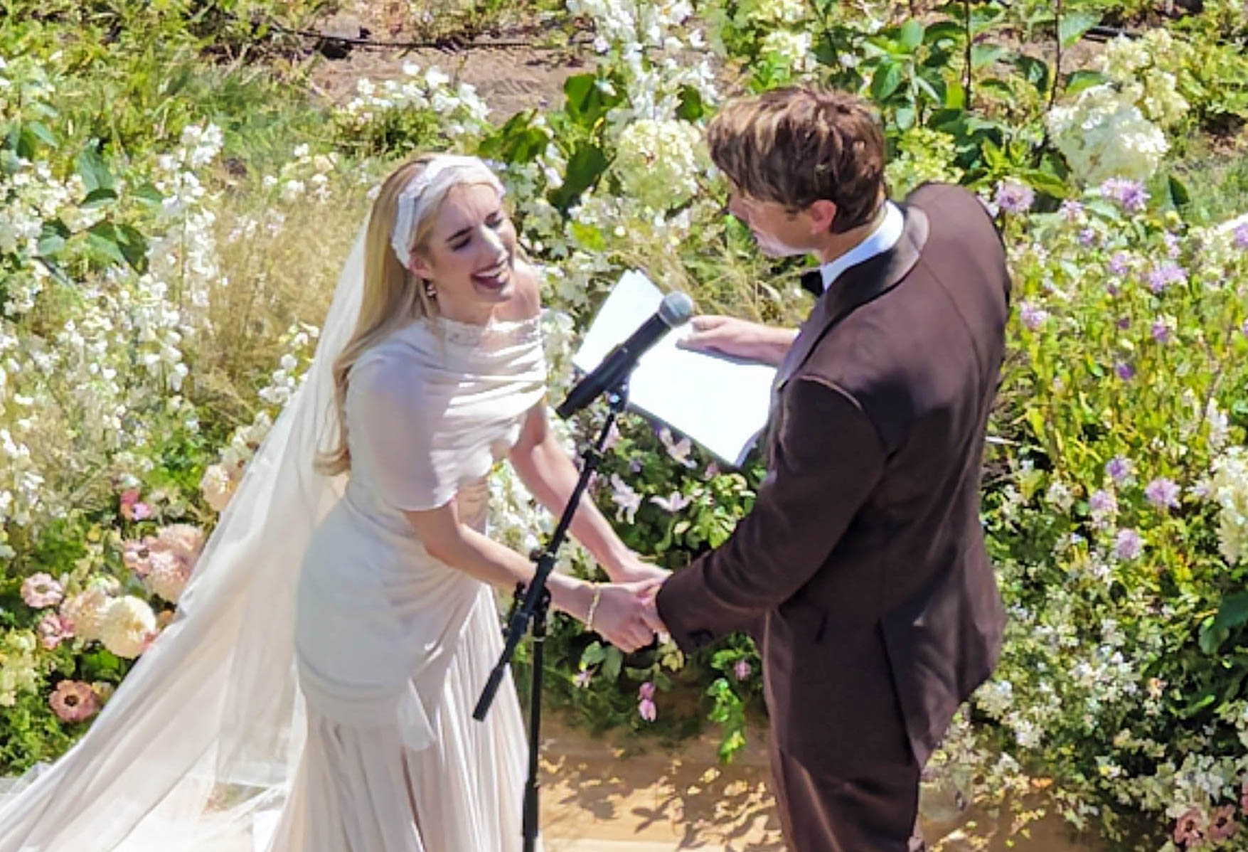 A bride in a white gown and veil laughs joyfully while holding hands with a groom in a brown suit at an outdoor ceremony surrounded by lush floral arrangements.