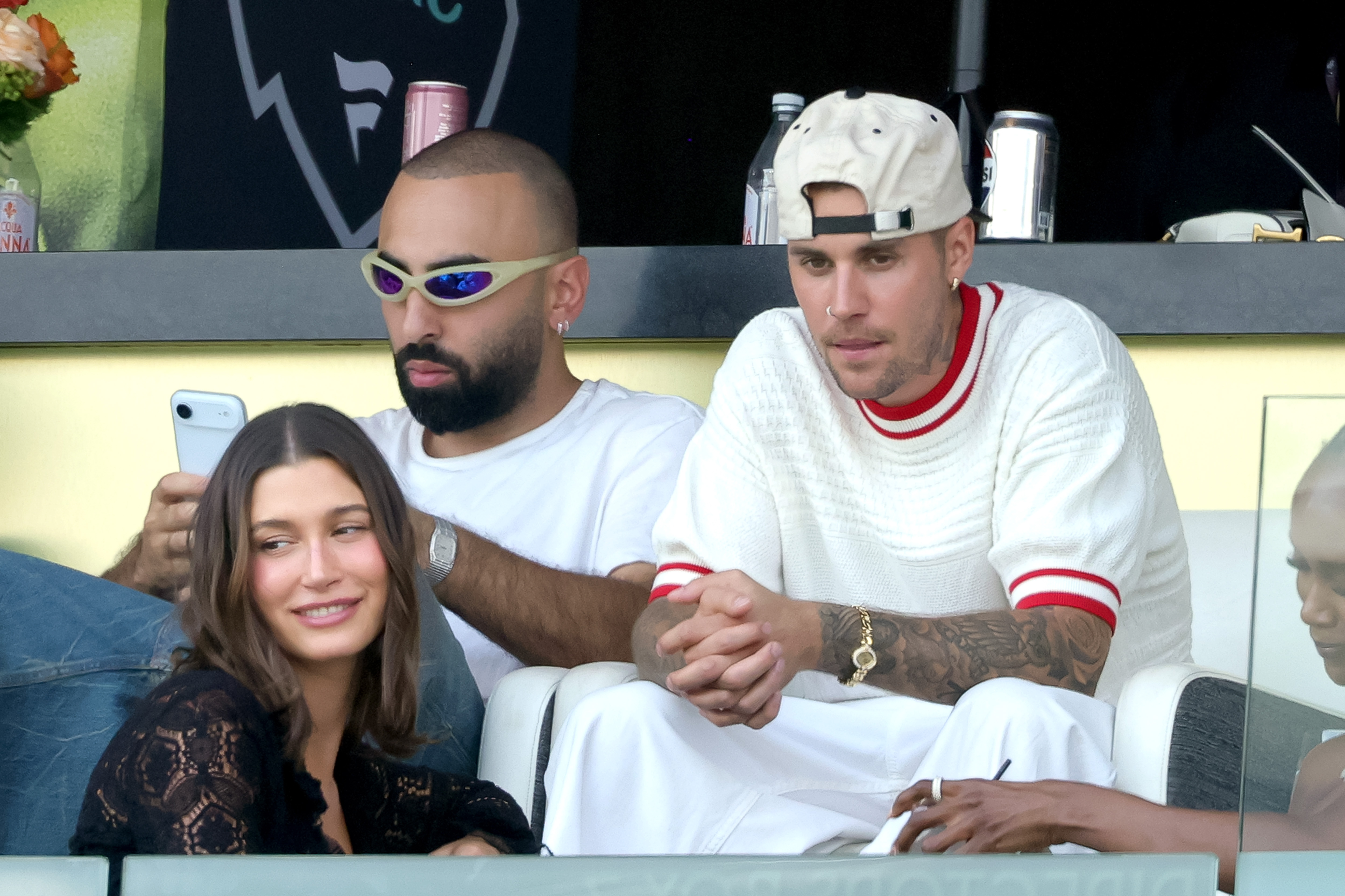 Woman in a black lace top and man in a white-and-red knit shirt holding hands in stadium seating, a bearded man with futuristic sunglasses seated between them.