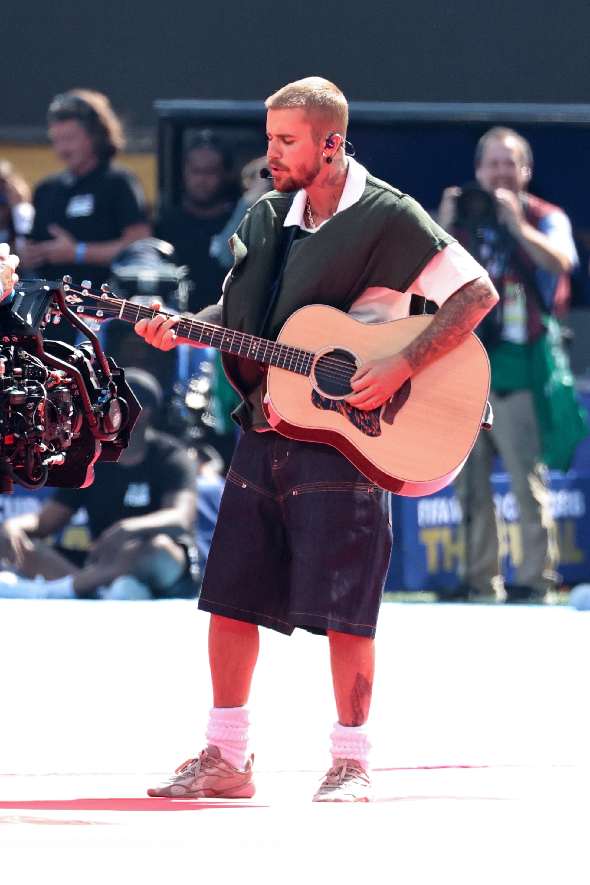 Man with short hair playing an acoustic guitar on a bright outdoor stage, wearing a dark green shirt, denim shorts, and white chunky socks.