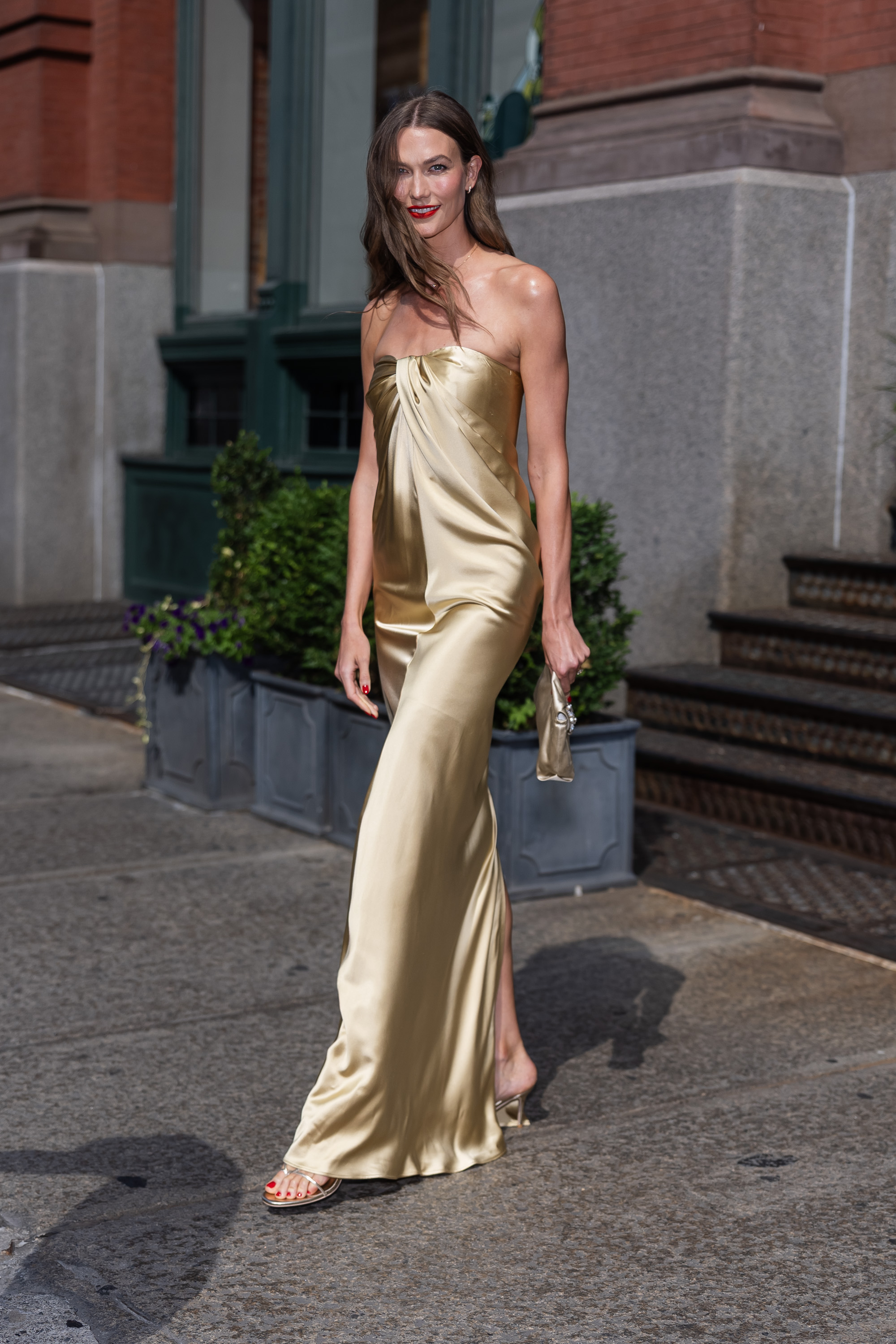 Click to open the image gallery Karlie Kloss walking on a SoHo sidewalk in a gold strapless satin floor-length gown and gold heeled sandals, holding a small clutch.
