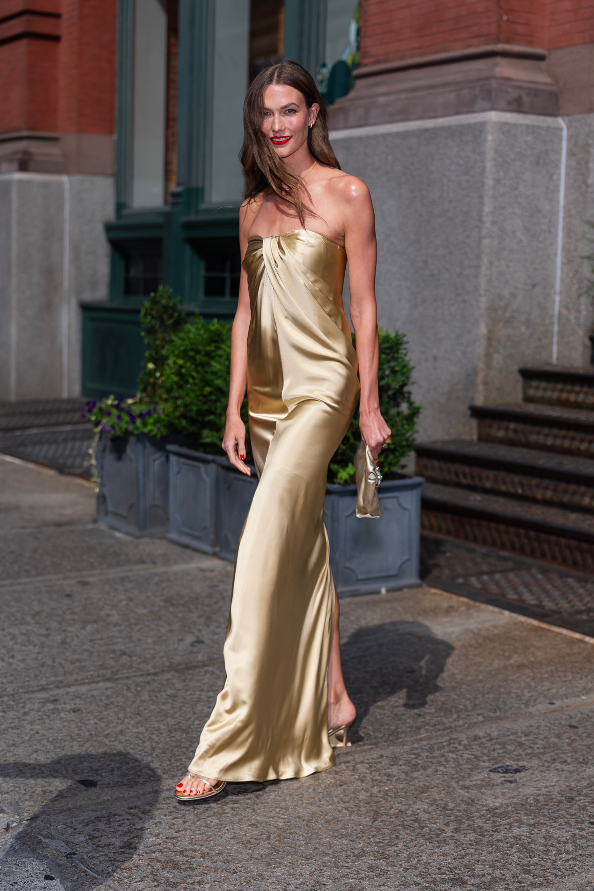 Karlie Kloss walking on a SoHo sidewalk in a gold strapless satin floor-length gown and gold heeled sandals, holding a small clutch.