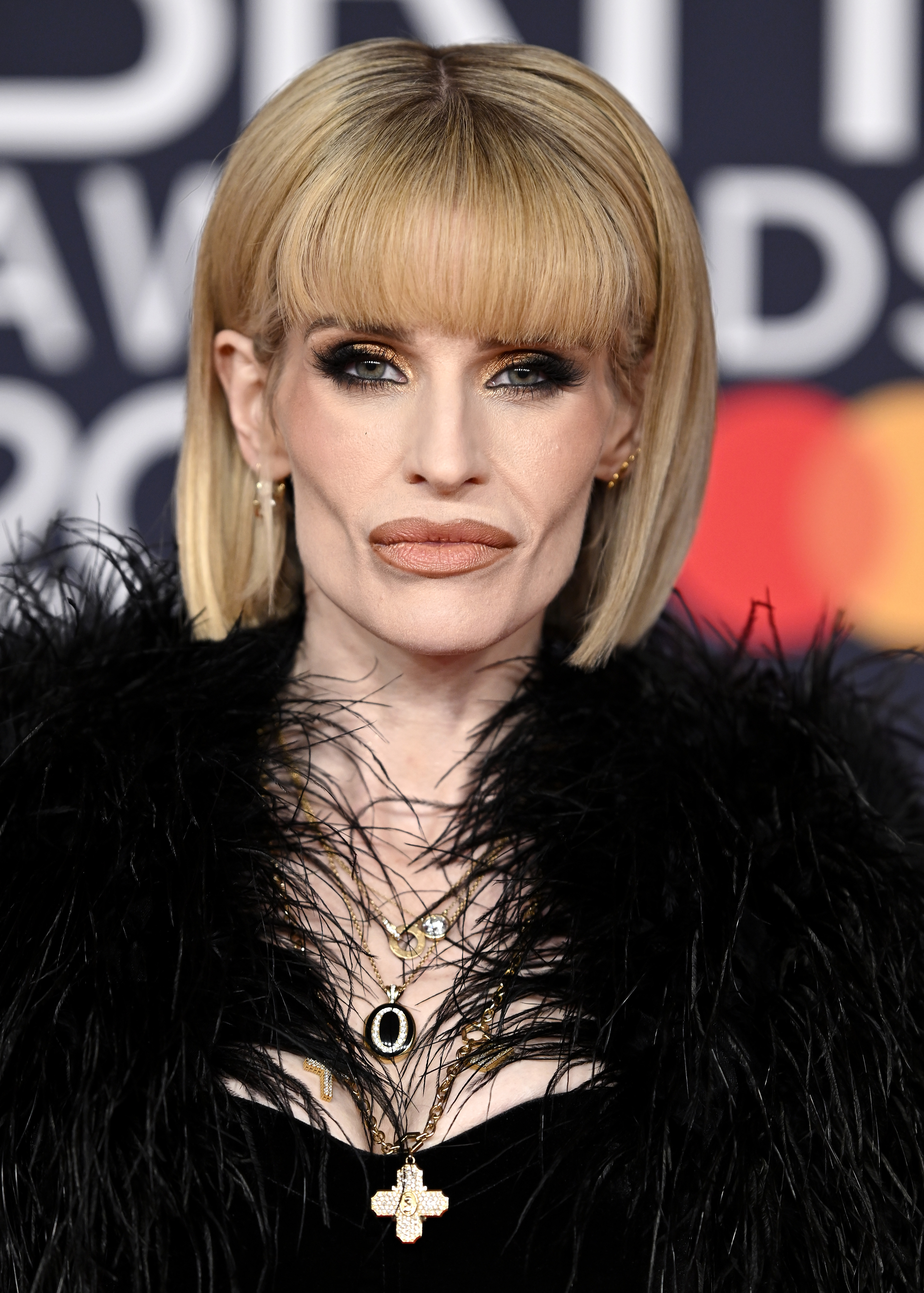 Kelly Osbourne in a black feathered outfit with gold layered necklaces and smoky eye makeup at the BRIT Awards 2026 in Manchester.