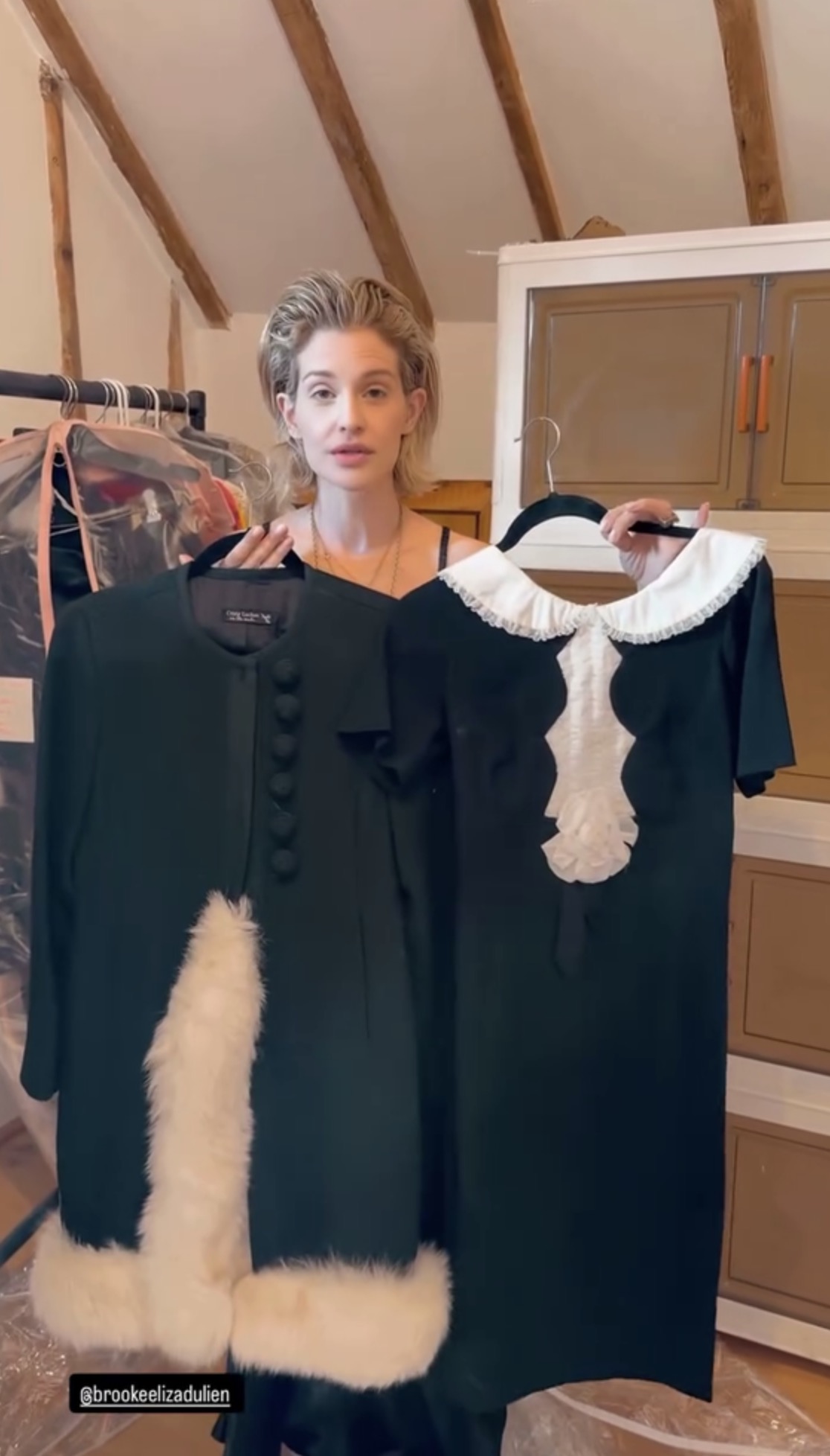 Makeup-free woman holding two black garments on hangers — a fur-trimmed coat and a white-collared dress — in a room with exposed wooden beams.