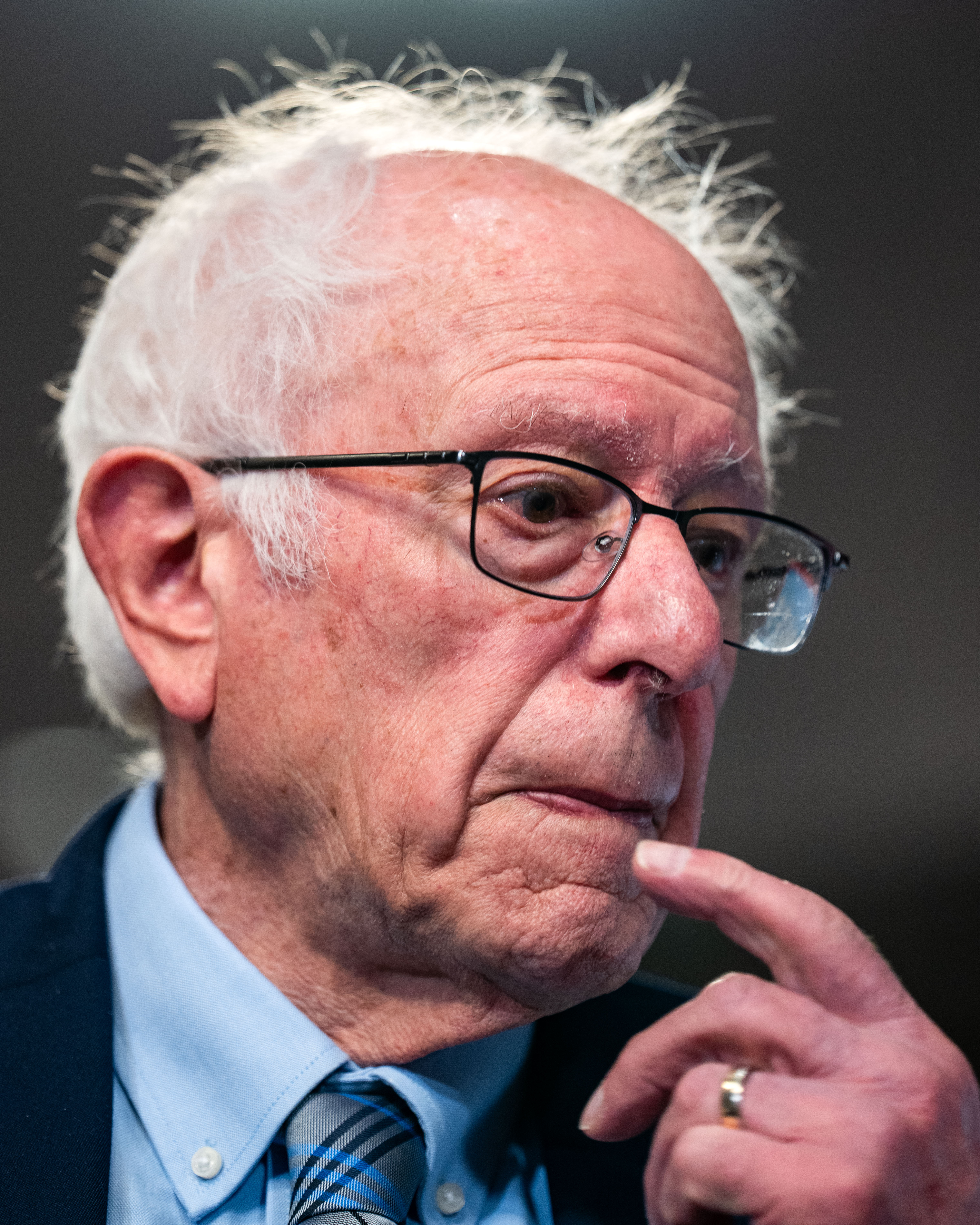 Sen. Bernie Sanders, an older white man with white hair and glasses, listens thoughtfully with his hand raised to his chin, wearing a blue shirt and striped tie.