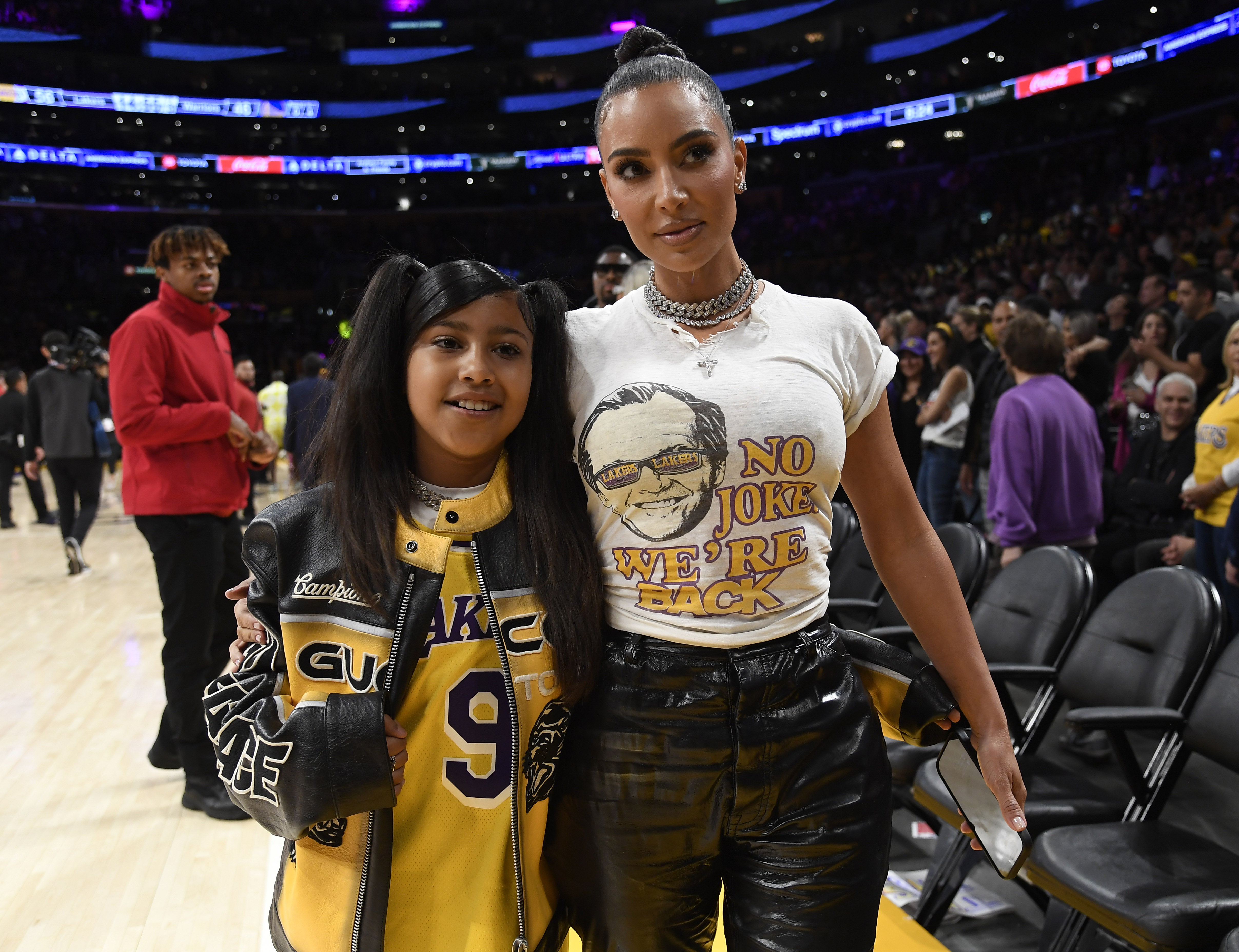 Kim Kardashian and daughter North West posing together courtside at a Los Angeles Lakers playoff game, North in a yellow Lakers jersey and Kim in a white graphic tee.
