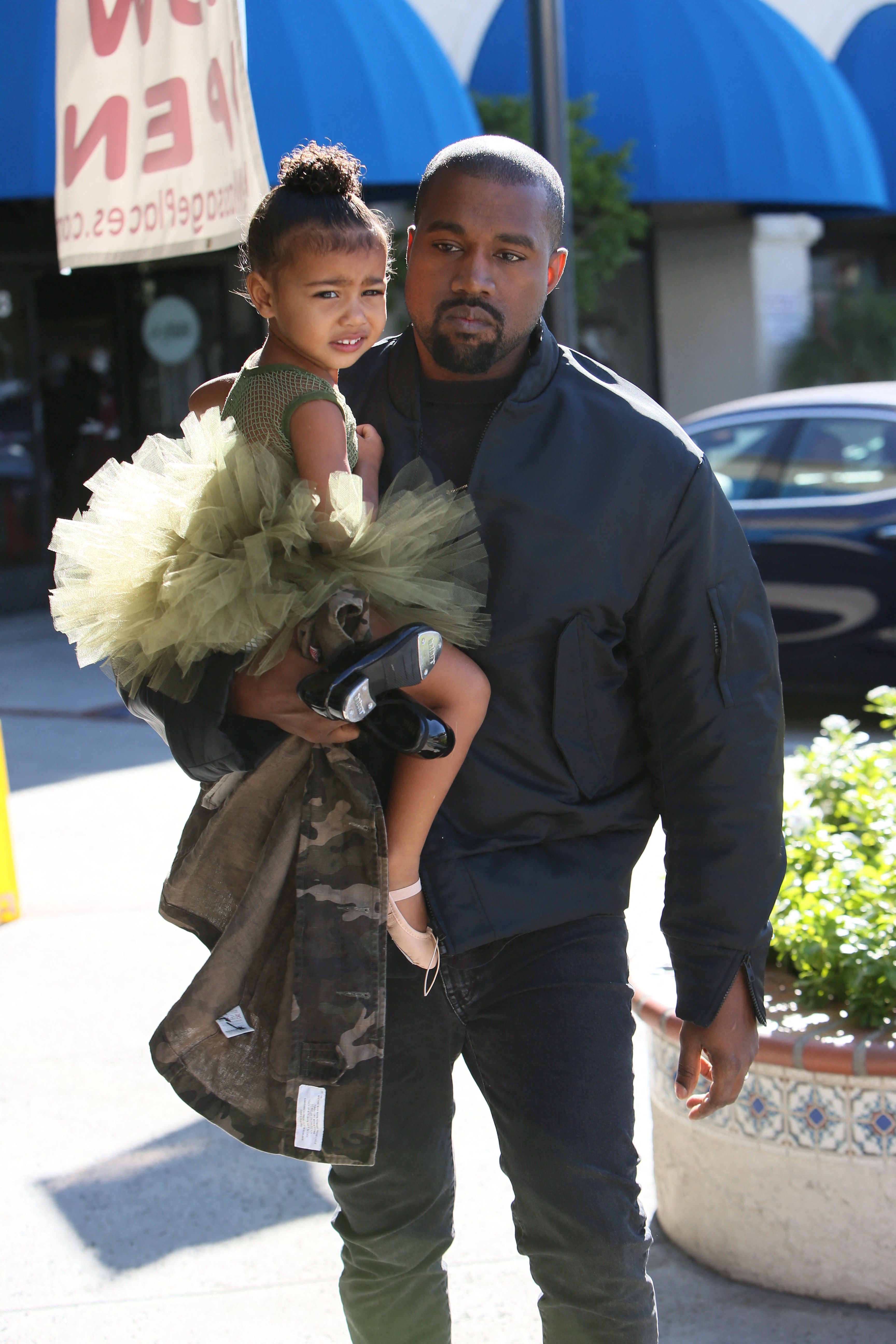 A man carrying a toddler-aged North West in a green tutu and camouflage jacket outside a building in Los Angeles.