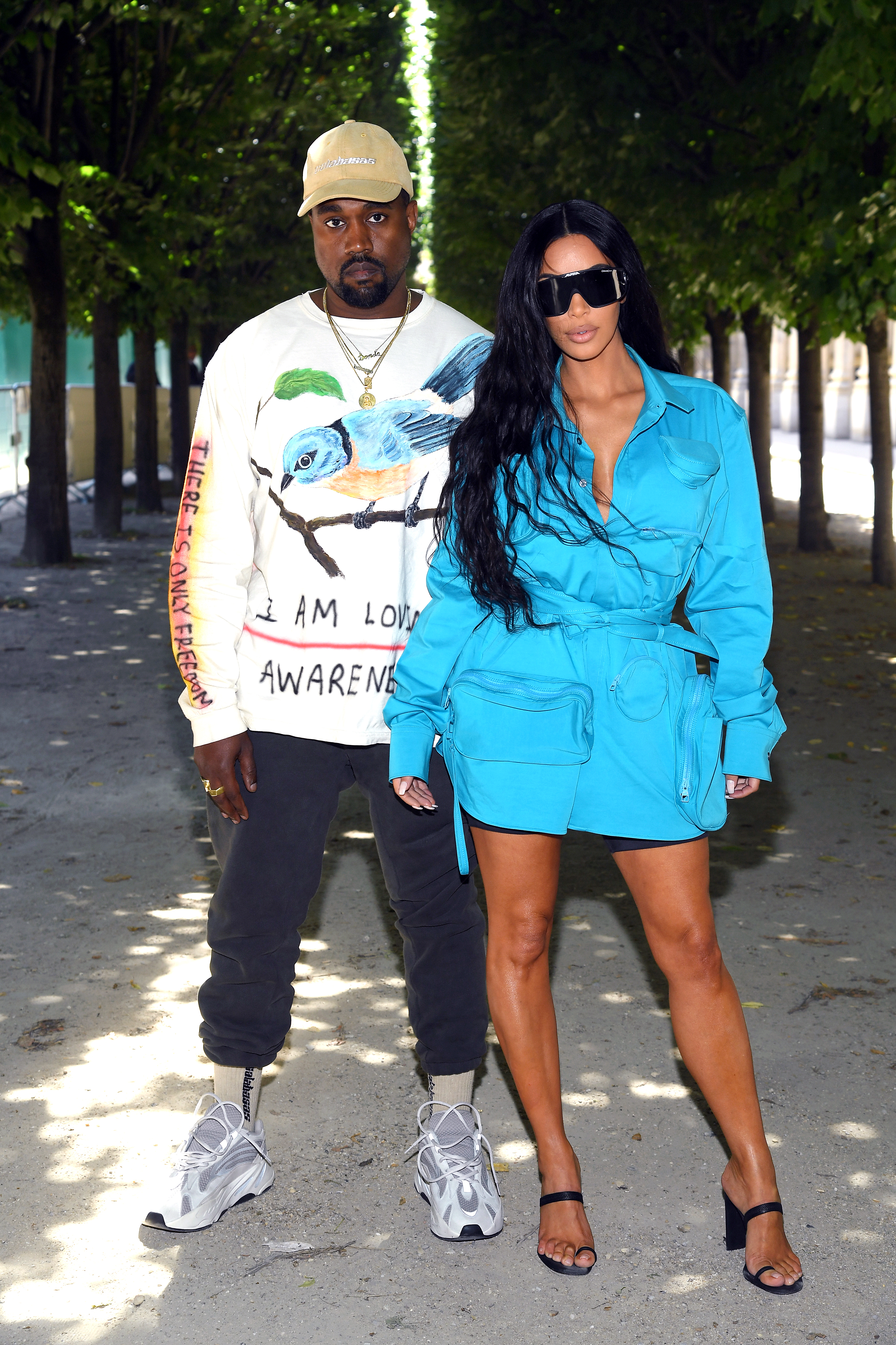 Kim Kardashian in a blue utility dress and Kanye West in a painted bird sweatshirt and cap posing outdoors at Paris Fashion Week.