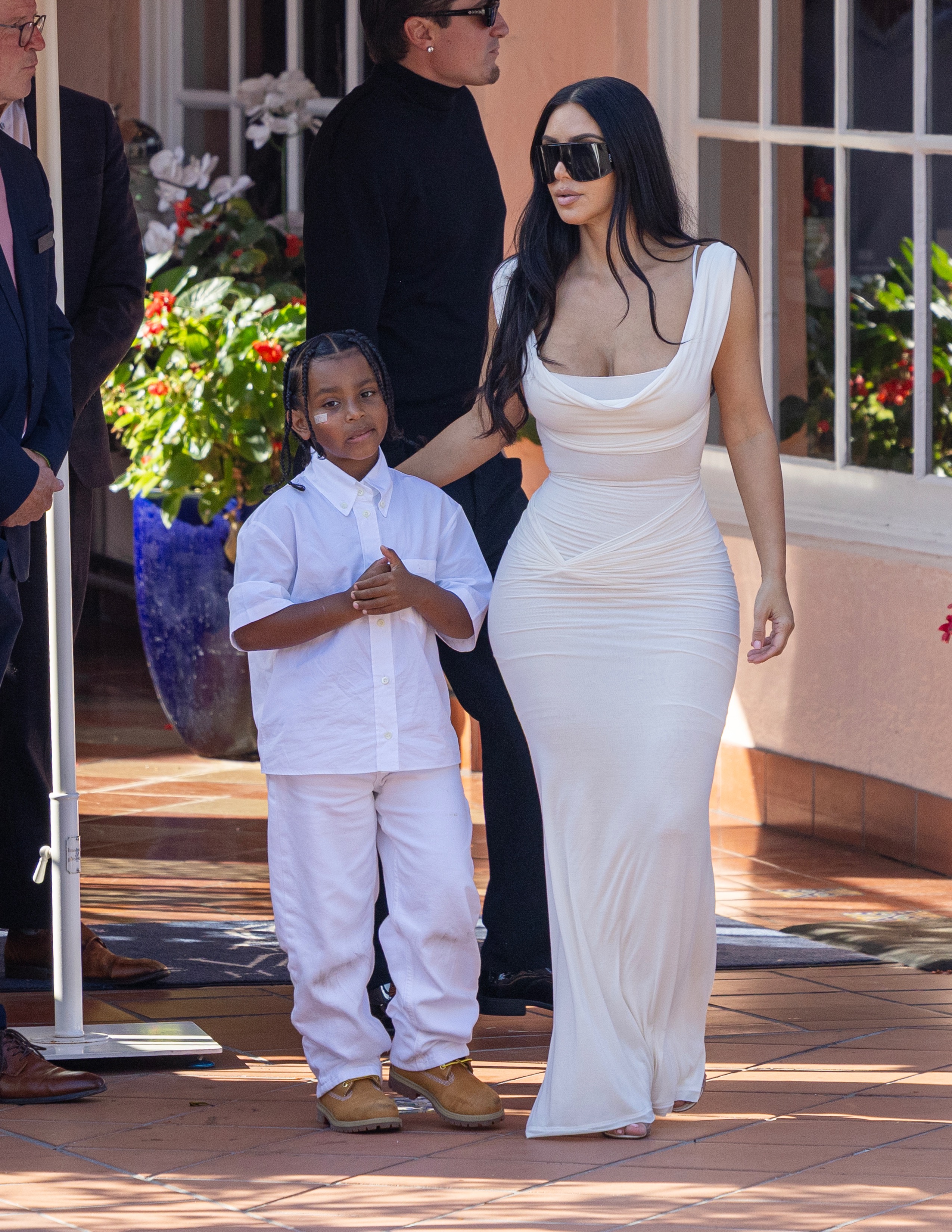 Woman in white sleeveless gown and large sunglasses walks with a young boy in white shirt and pants outside a pink hotel.