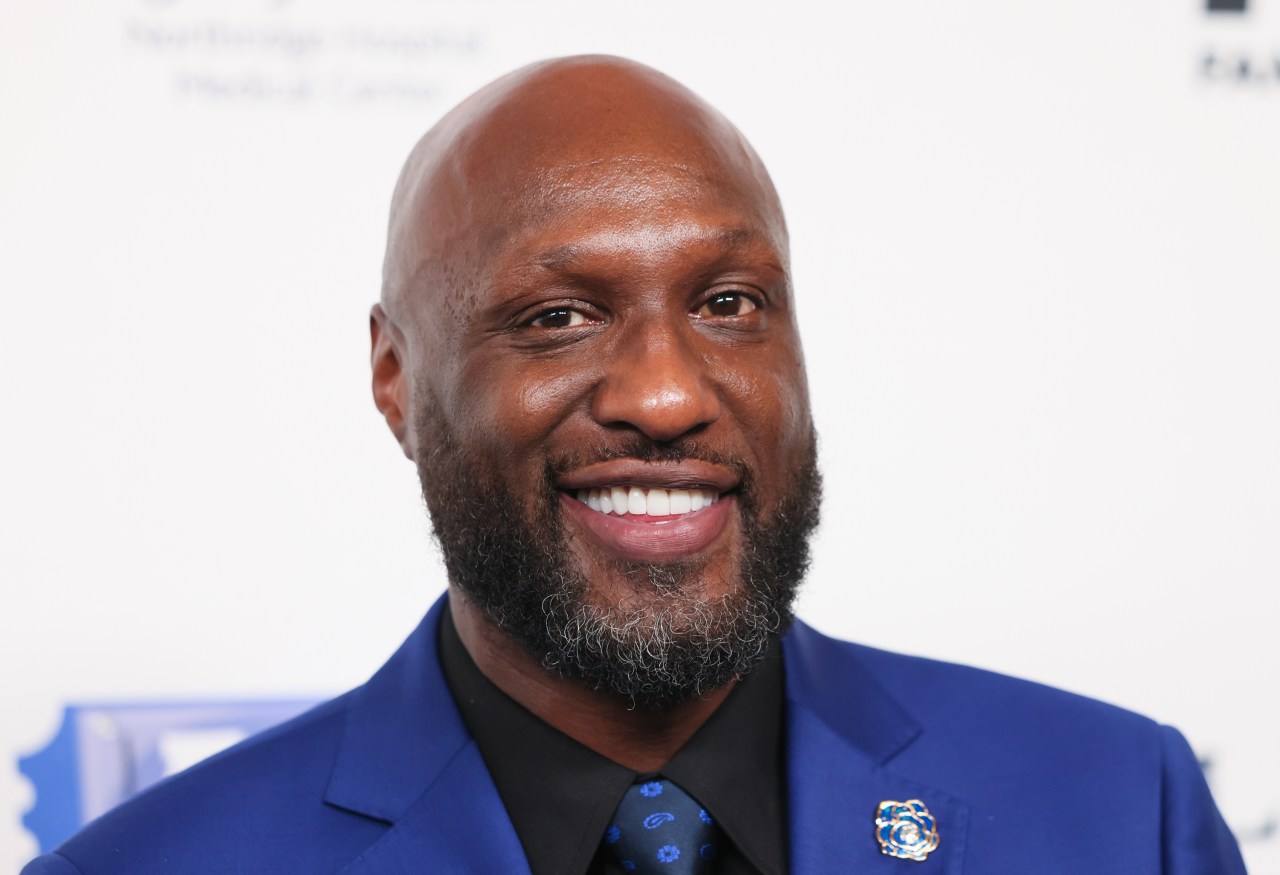 Lamar Odom smiling in a blue suit with a black shirt and blue tie at a red carpet event in Beverly Hills.