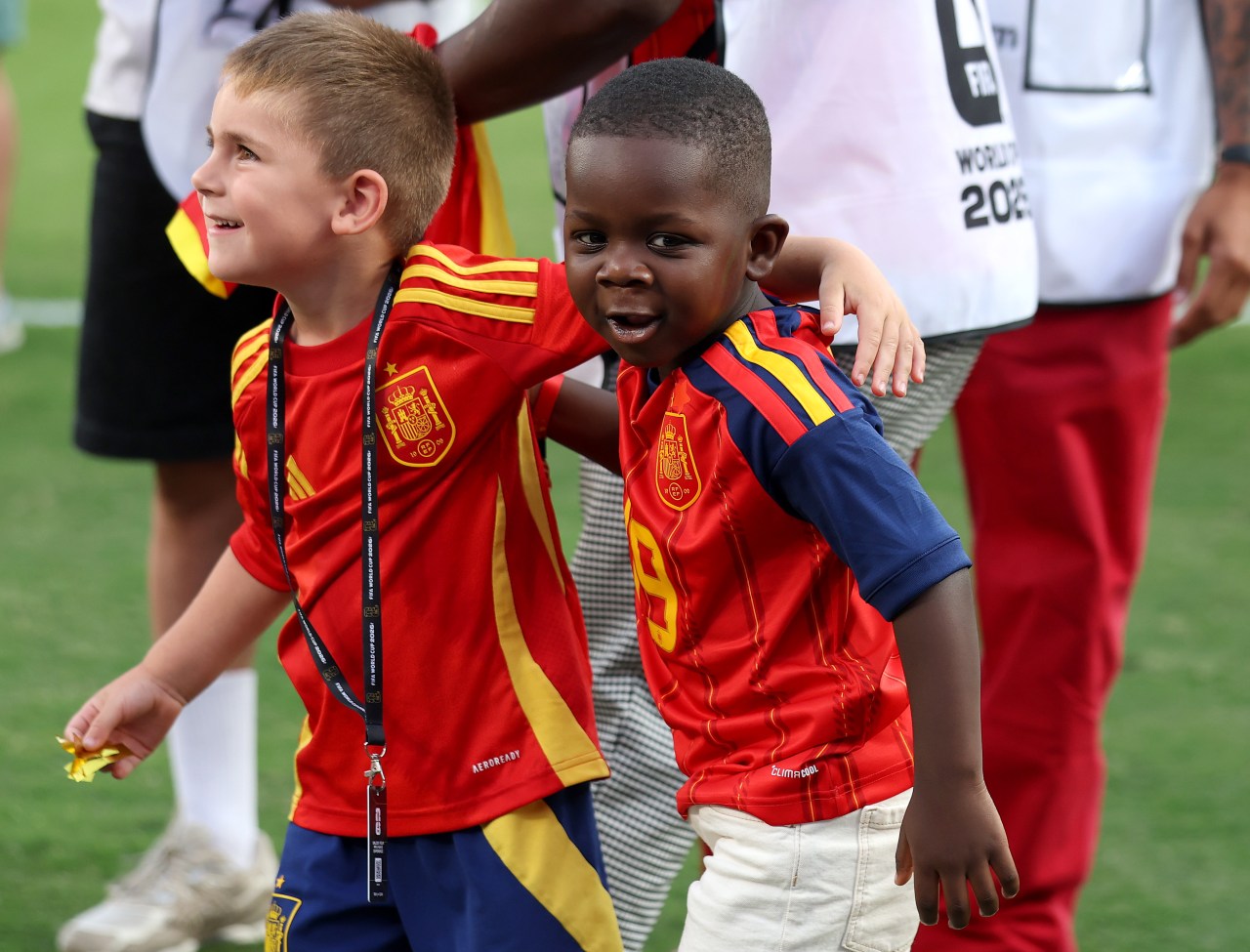 Two young boys wearing Spain's red number 19 jerseys celebrate on a football pitch, one smiling broadly and the other with his arm around the other's shoulder.