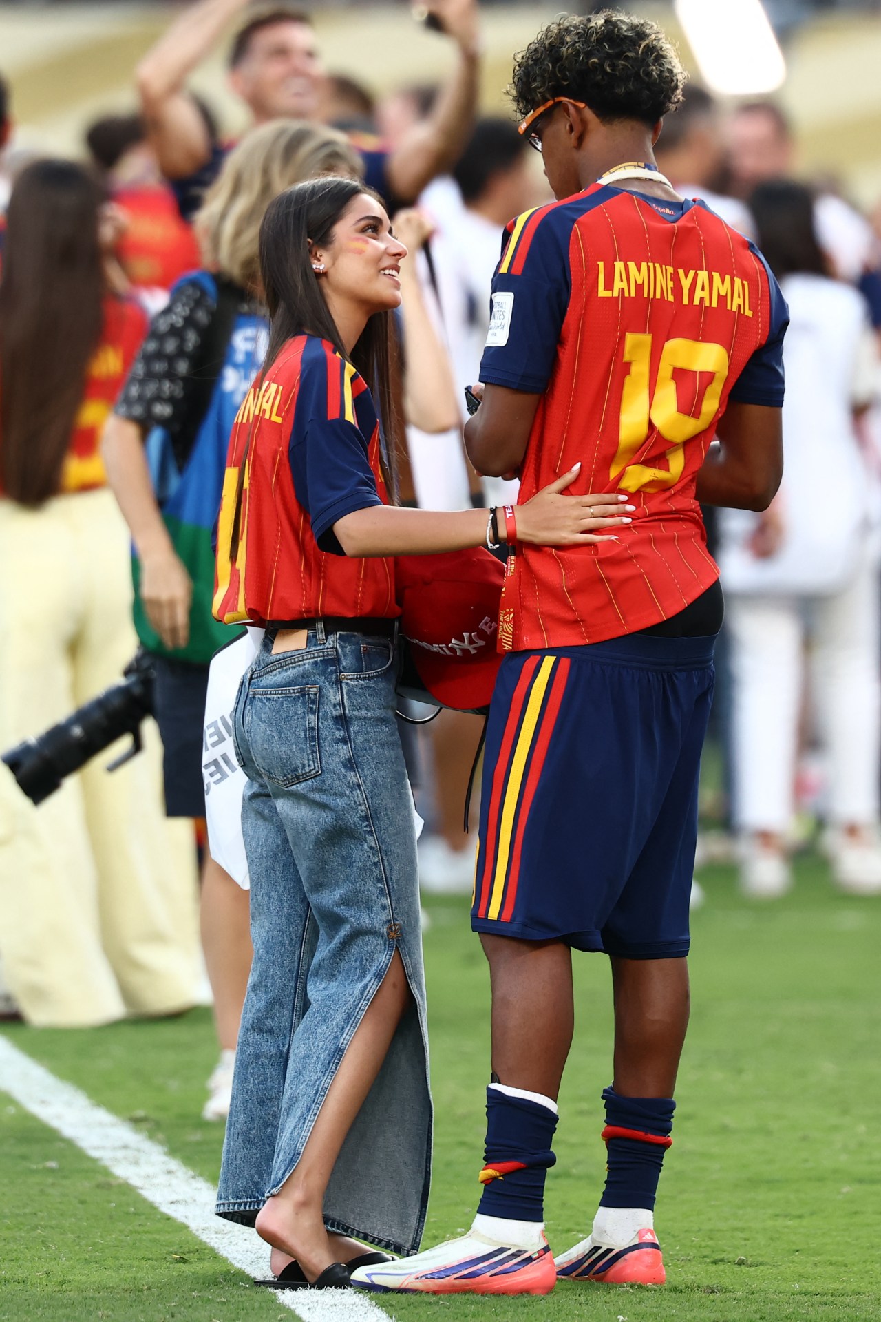 A young woman in a Spain Lamine Yamal jersey and jeans looks up at a footballer wearing a number 19 Spain kit on a football pitch after a match.