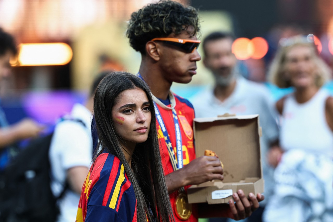 Young woman with red and yellow face paint standing next to a young man in a Spain jersey holding an open pizza box after a football match.