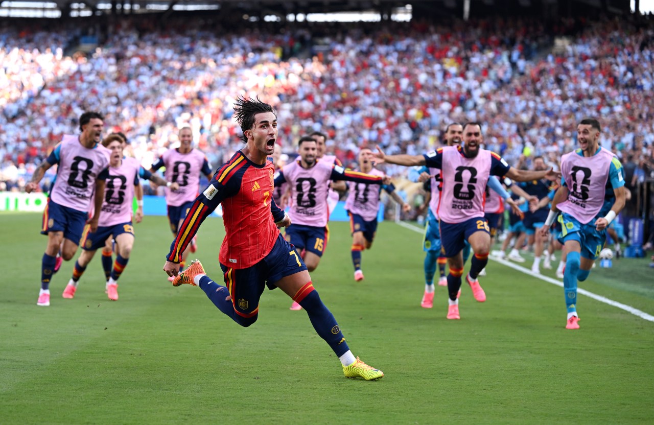 Ferran Torres in Spain's red kit leaping in celebration as teammates in pink training bibs sprint toward him on a football pitch.