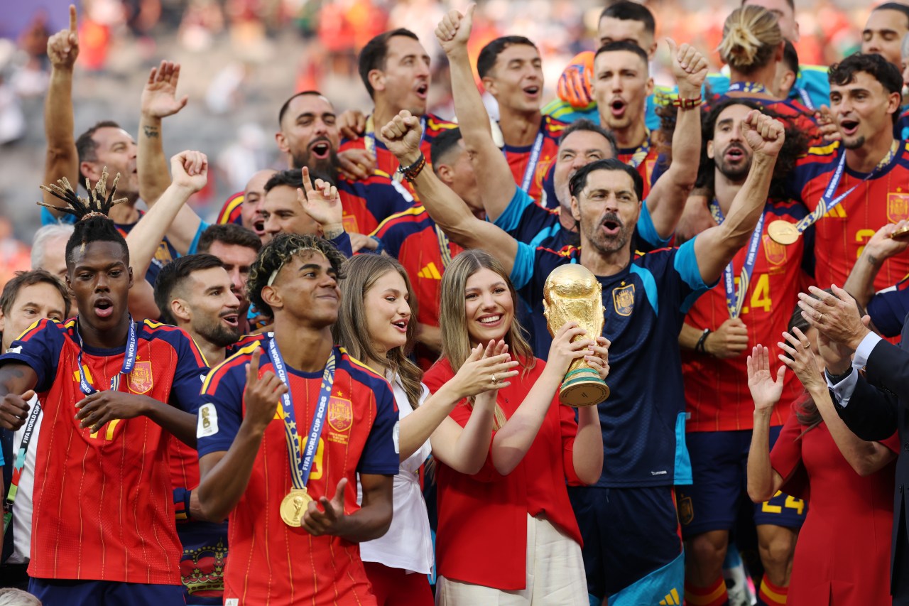 Princess Sofía holding the golden FIFA World Cup trophy surrounded by celebrating Spain players and Princess Leonor at the post-match award ceremony.