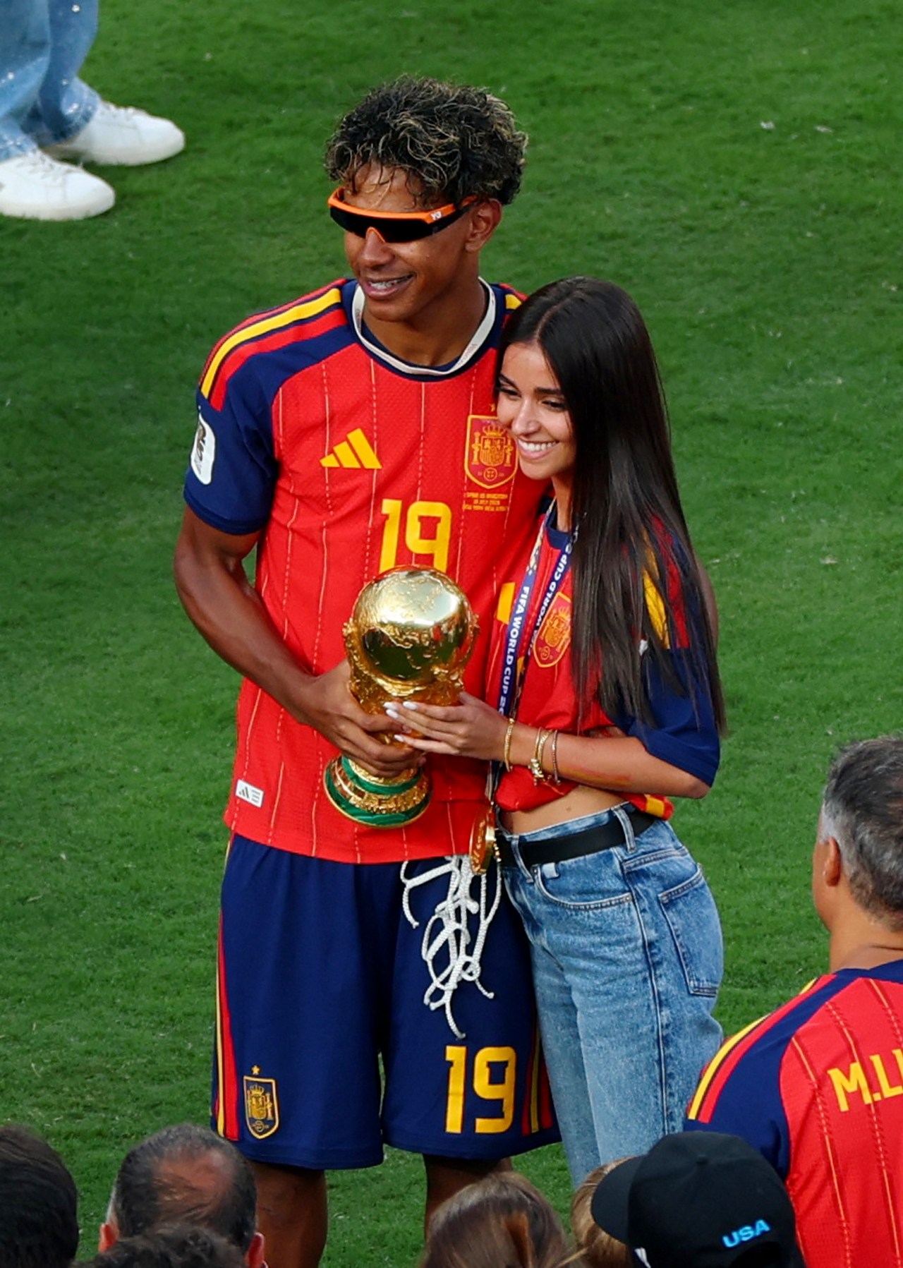 Young male footballer in Spain No. 19 red kit and orange-banded sunglasses holds the FIFA World Cup trophy alongside a smiling young woman in a Spain jersey on the pitch.