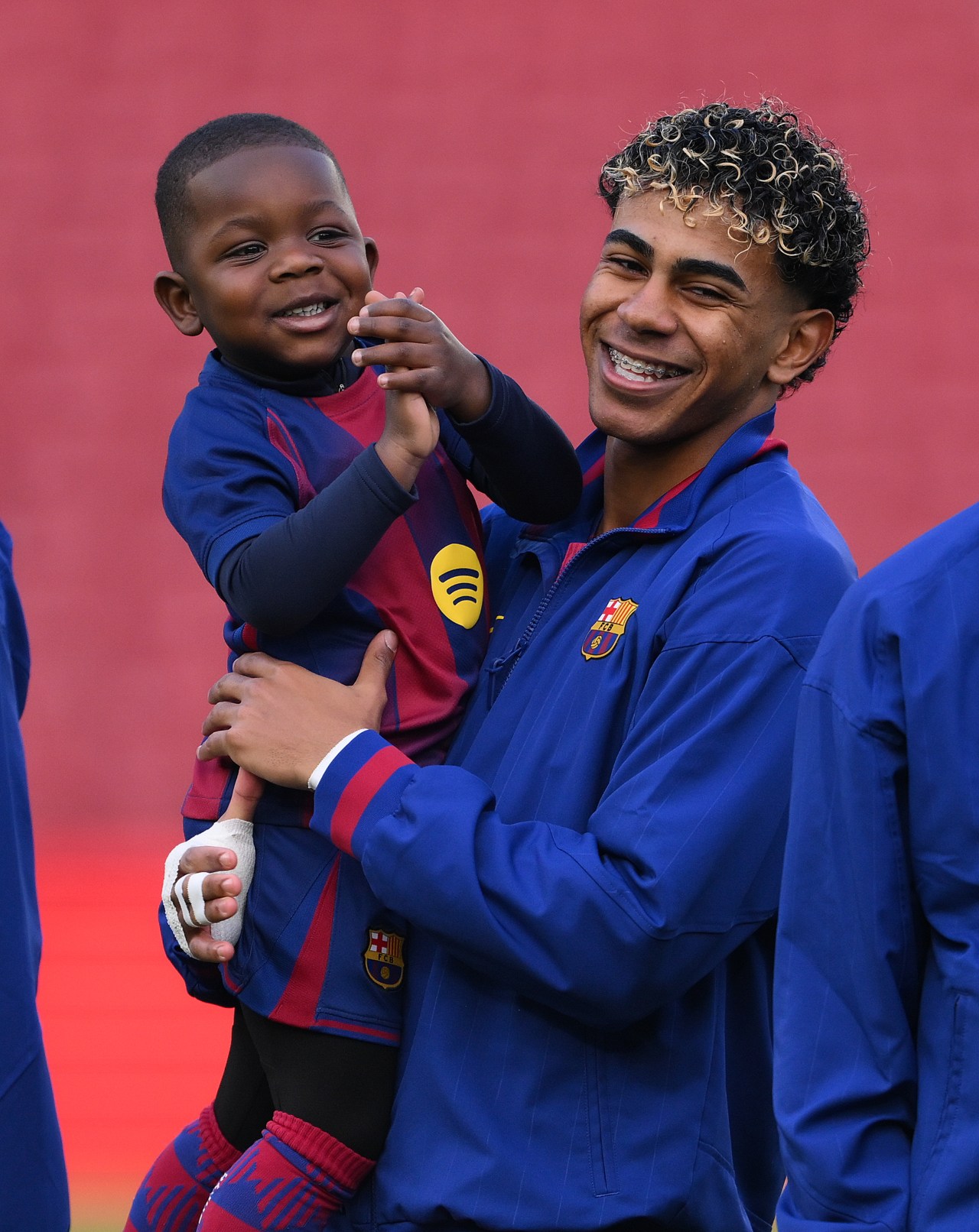 A young FC Barcelona player with curly hair holds a smiling toddler dressed in a Barcelona kit on a football pitch.