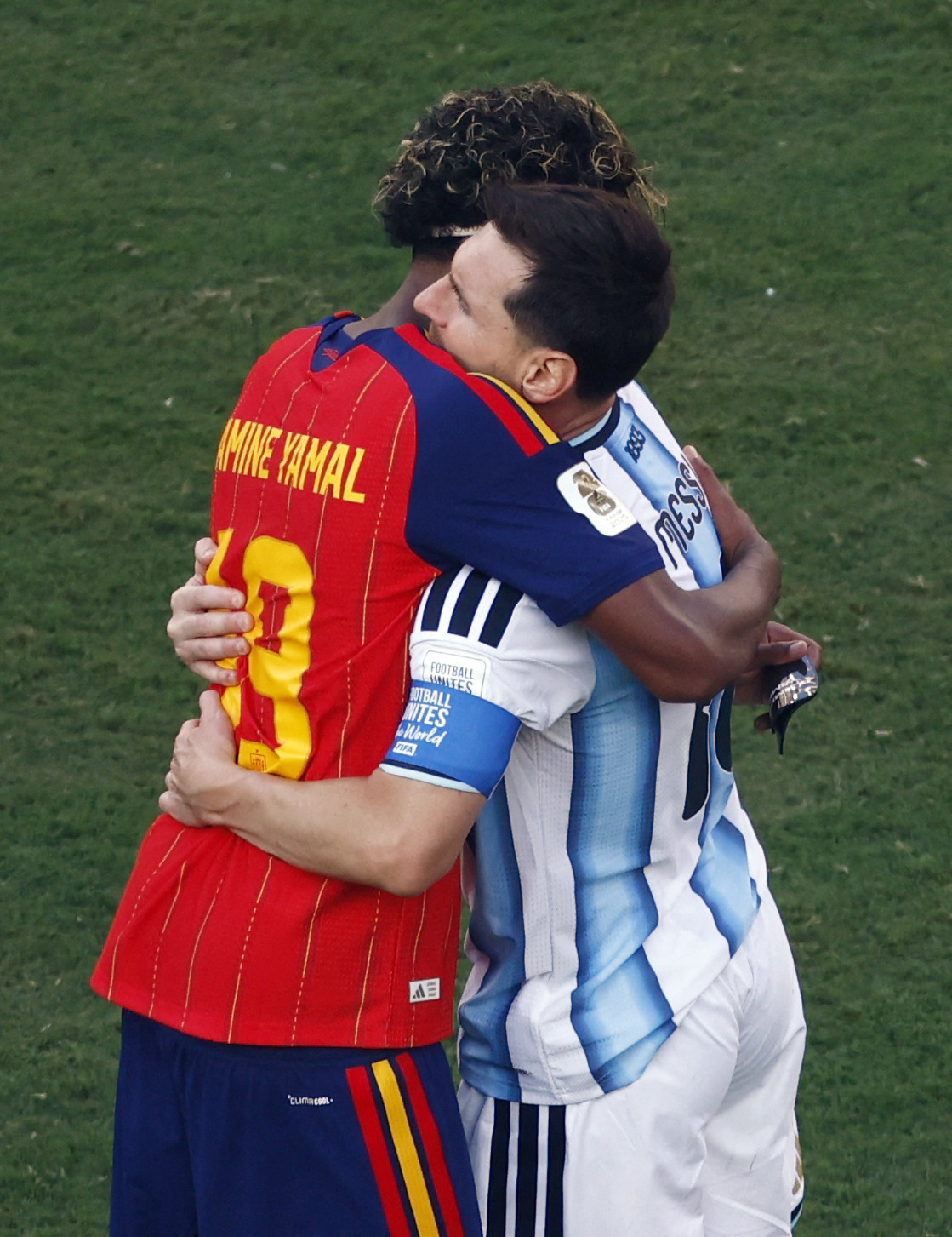 Lamine Yamal in Spain's red kit hugging Messi in Argentina's light blue and white jersey on a grass pitch after a match.