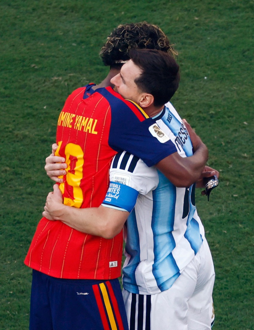 Lamine Yamal in Spain's red kit hugging Messi in Argentina's light blue and white jersey on a grass pitch after a match.