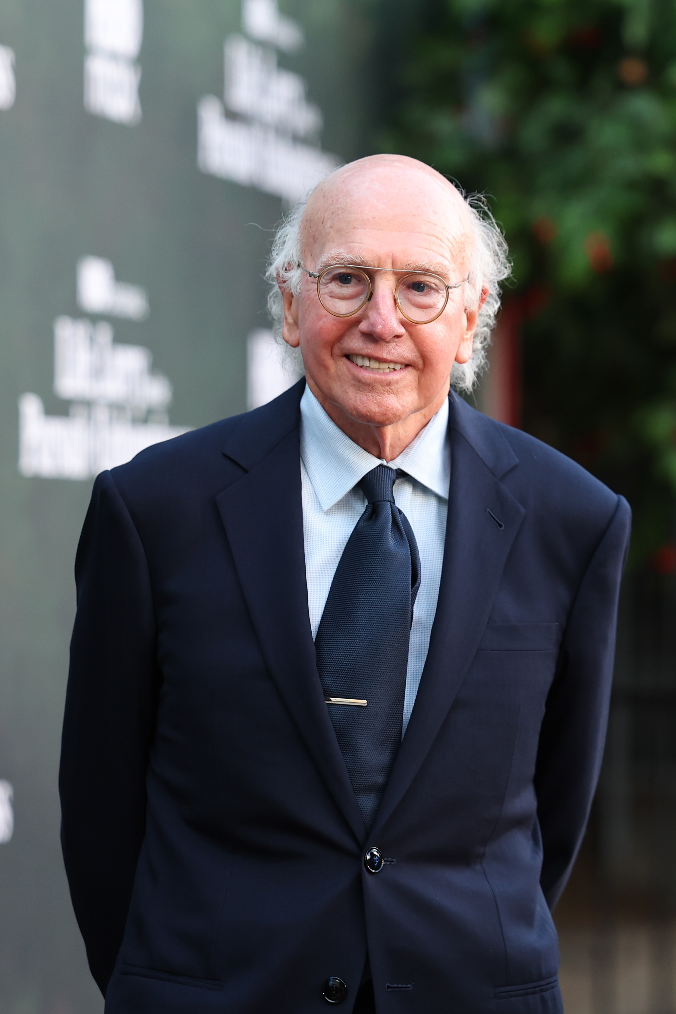 Larry David smiling in a dark suit and tie at a red carpet event, hands in pockets, in front of a branded backdrop.