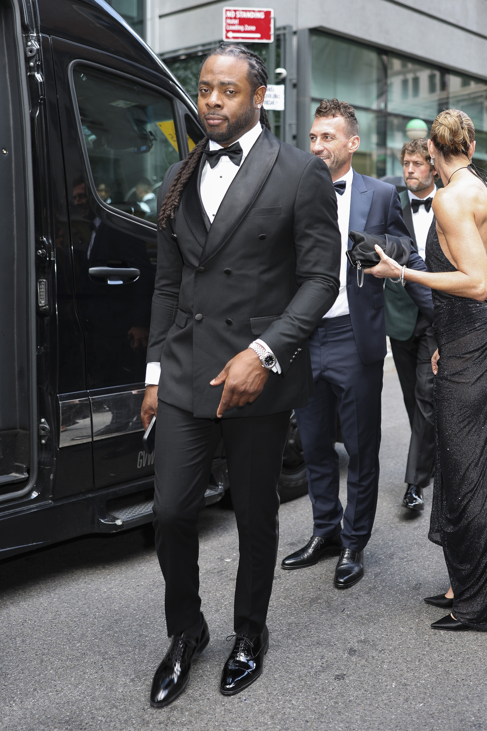 Richard Sherman in a black double-breasted tuxedo with bow tie and long braids, standing outside a black van on a city street.