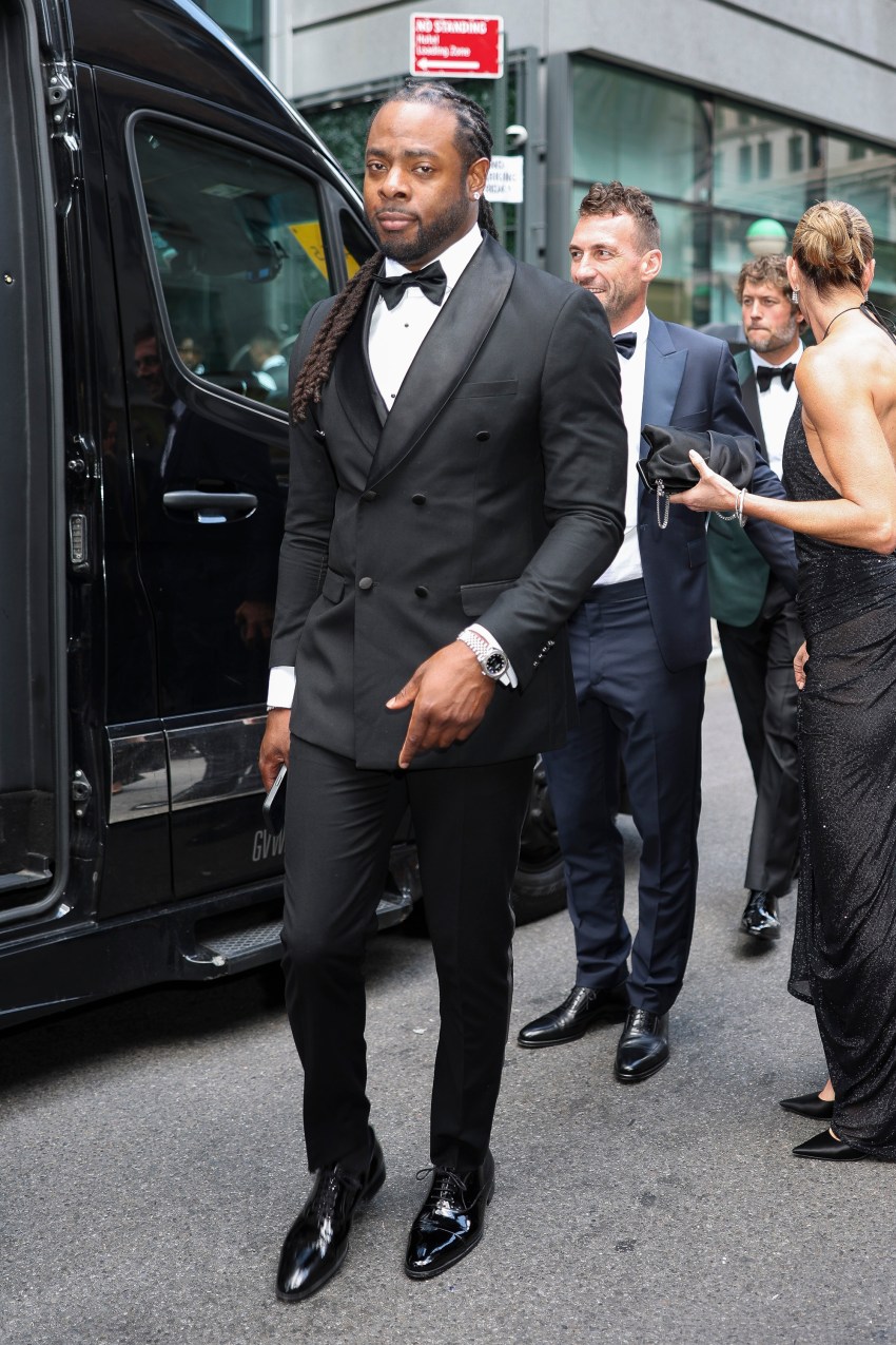Richard Sherman in a black double-breasted tuxedo with bow tie and long braids, standing outside a black van on a city street.
