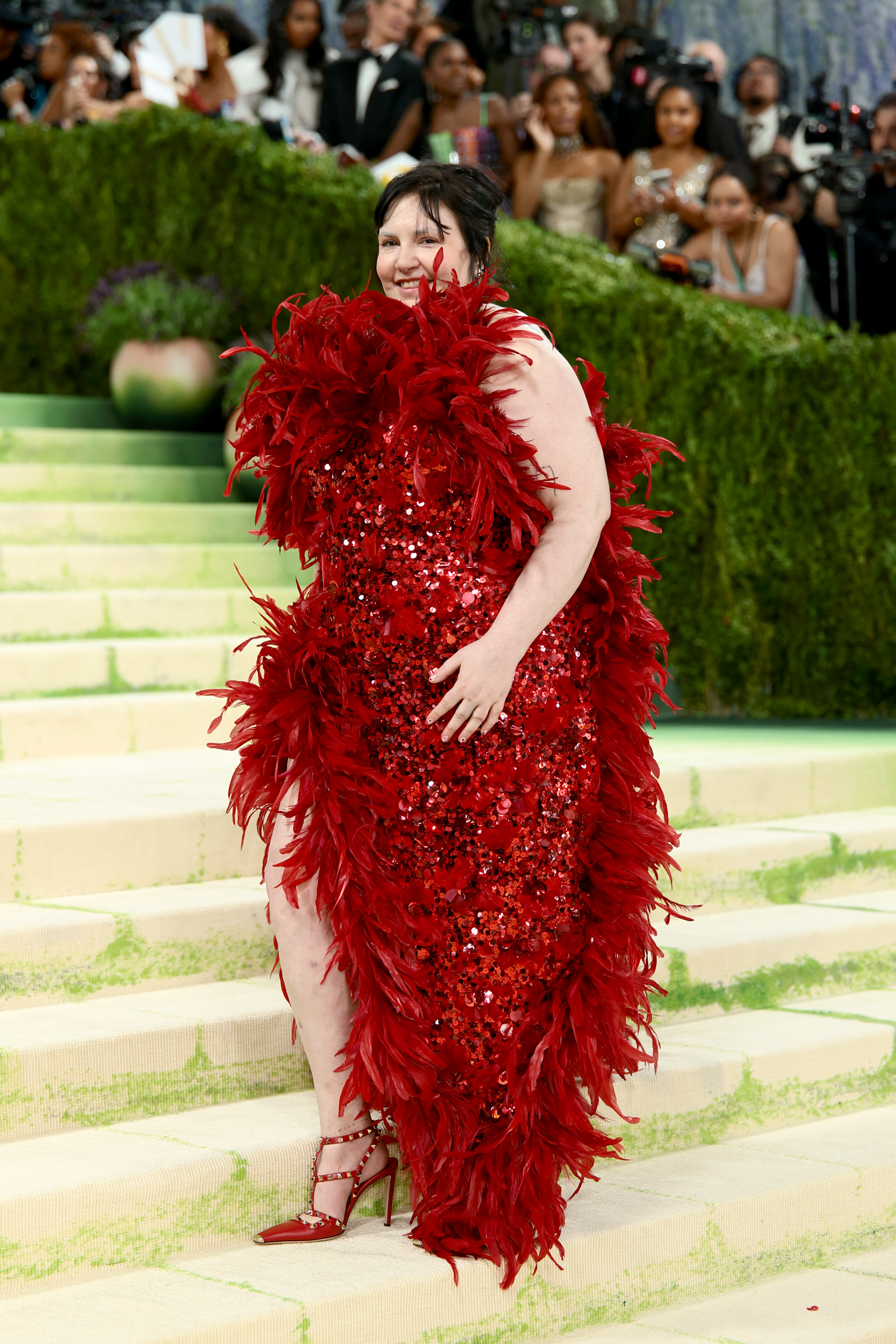 Lena Dunham smiling on stone steps in a red sequined dress covered in red feathers, wearing red strappy heels.