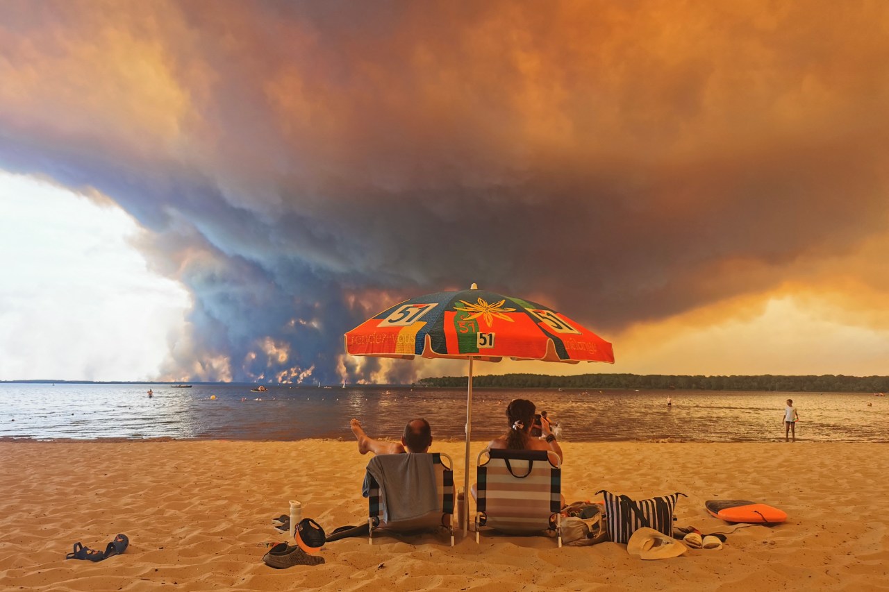 Two people sit in beach chairs under a colorful parasol on a sandy beach while enormous dark smoke clouds from a wildfire billow across the sky and lake behind them.