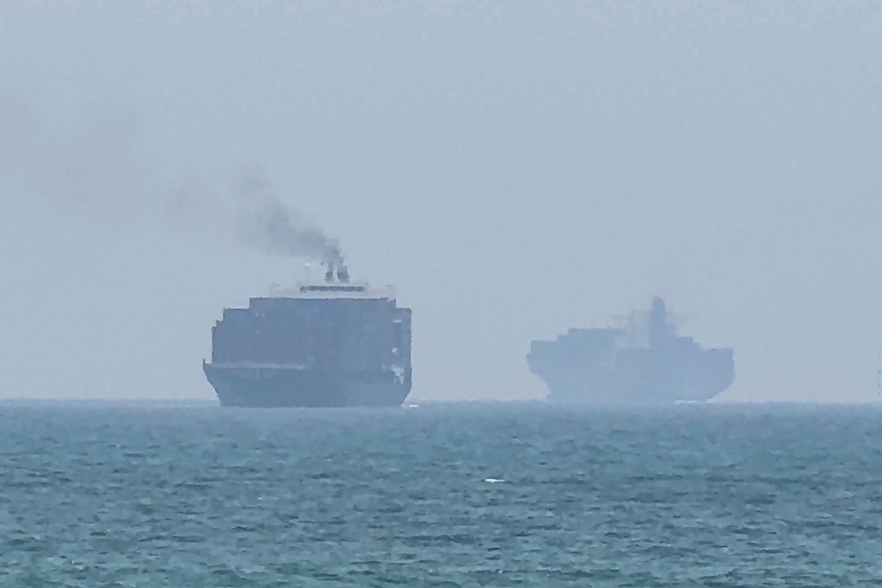 Two large cargo ships visible through haze on open sea near the Strait of Hormuz, with smoke rising from one vessel.