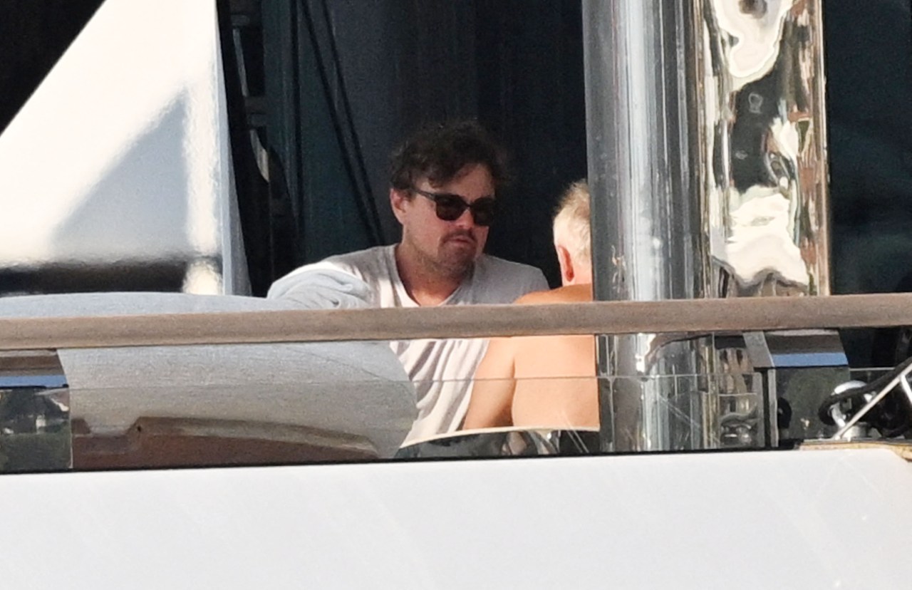 A man with dark curly hair and sunglasses wearing a gray shirt sits on the deck of a yacht, talking with another person.