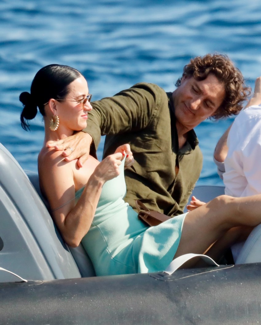 A woman in a light blue strapless outfit sits on a boat while a curly-haired man in an olive shirt reaches over to apply sunscreen to her shoulder.