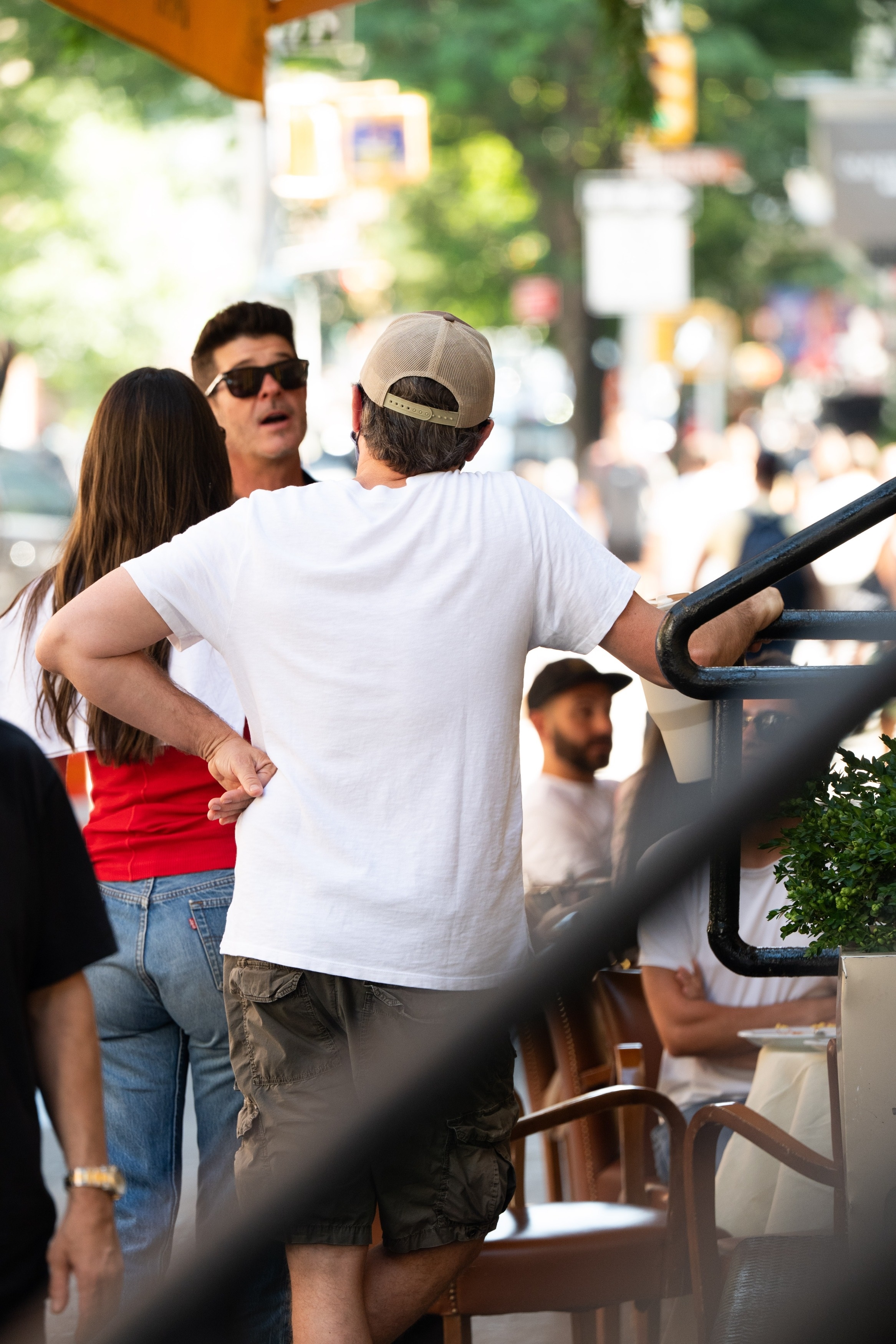 A man in a white t-shirt and backwards cap stands with his back to the camera speaking with a brunette woman and another man wearing sunglasses outside a restaurant.