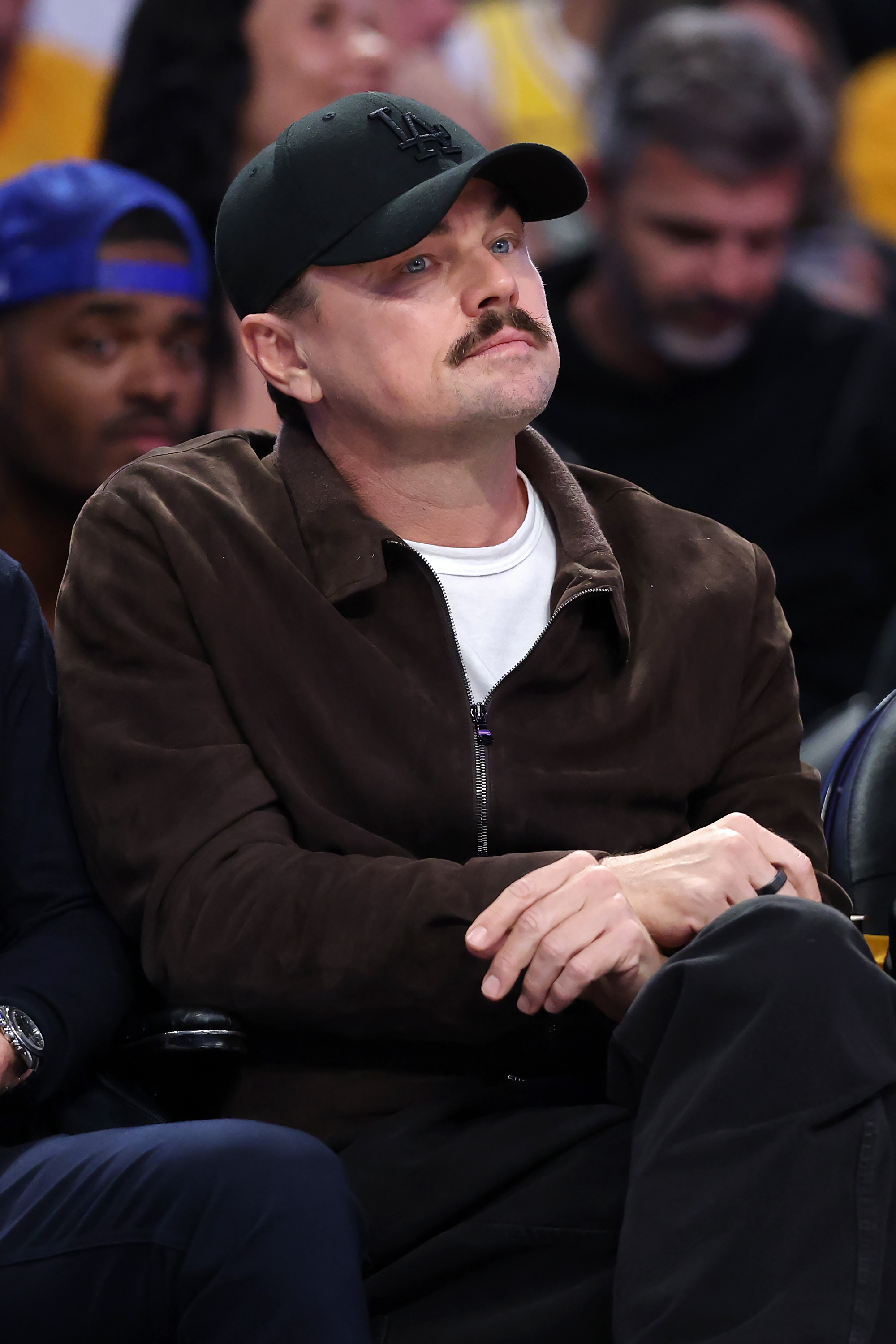 Man wearing a black LA cap, mustache, and brown zip-up jacket sits courtside at an NBA game with hands clasped, looking upward.