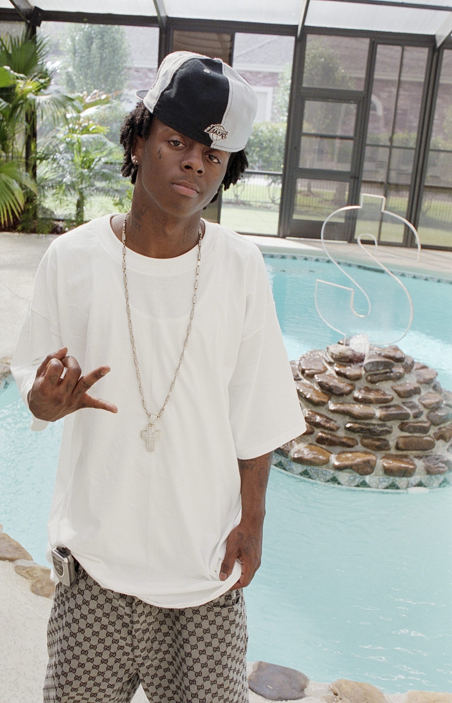 Young Lil' Wayne in a white tee, Lakers cap, and Gucci pants standing in front of an indoor pool, making a hand gesture.