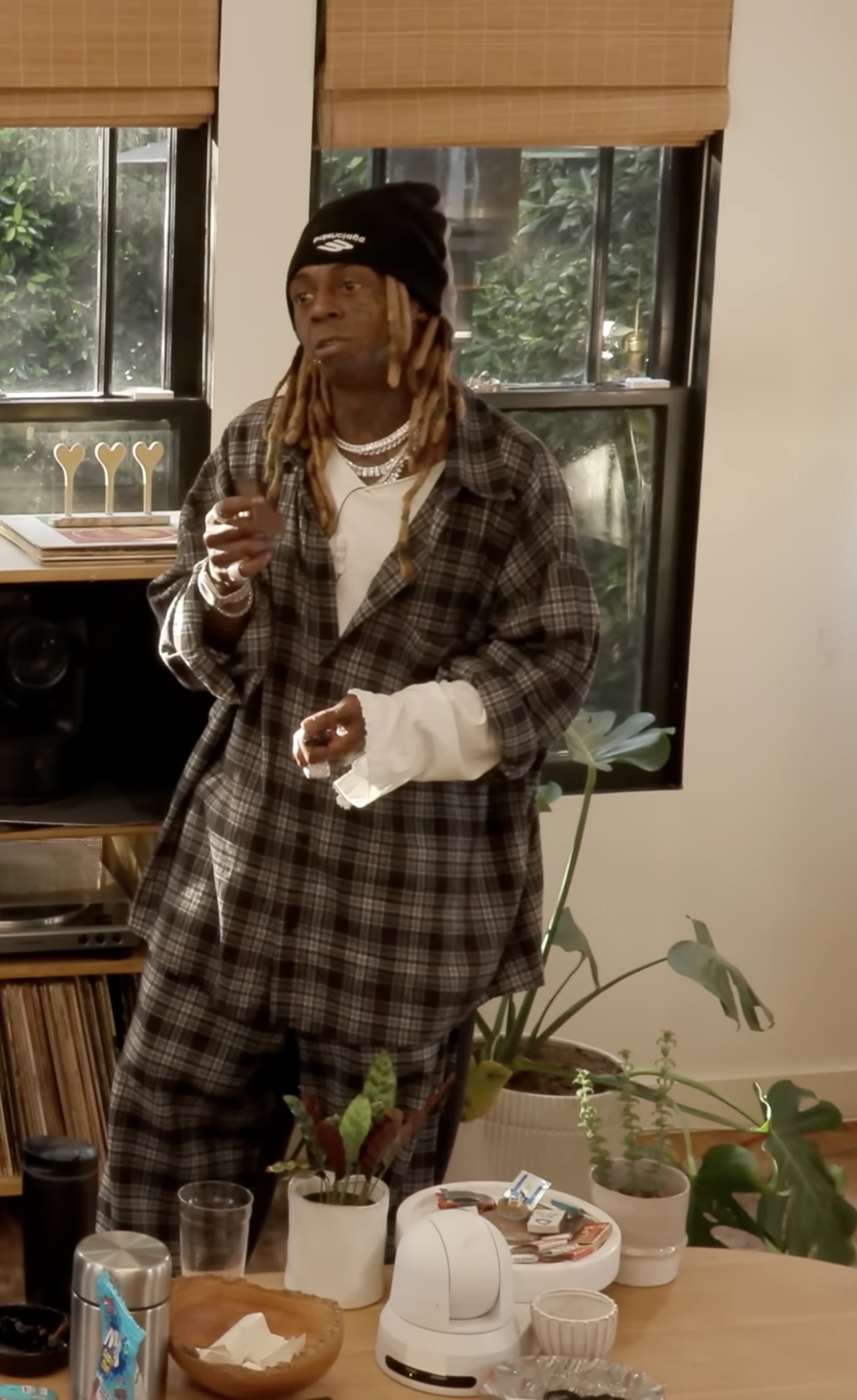 Lil' Wayne with long blonde locs and a black beanie, dressed in plaid, standing indoors near houseplants and a shelf of vinyl records.