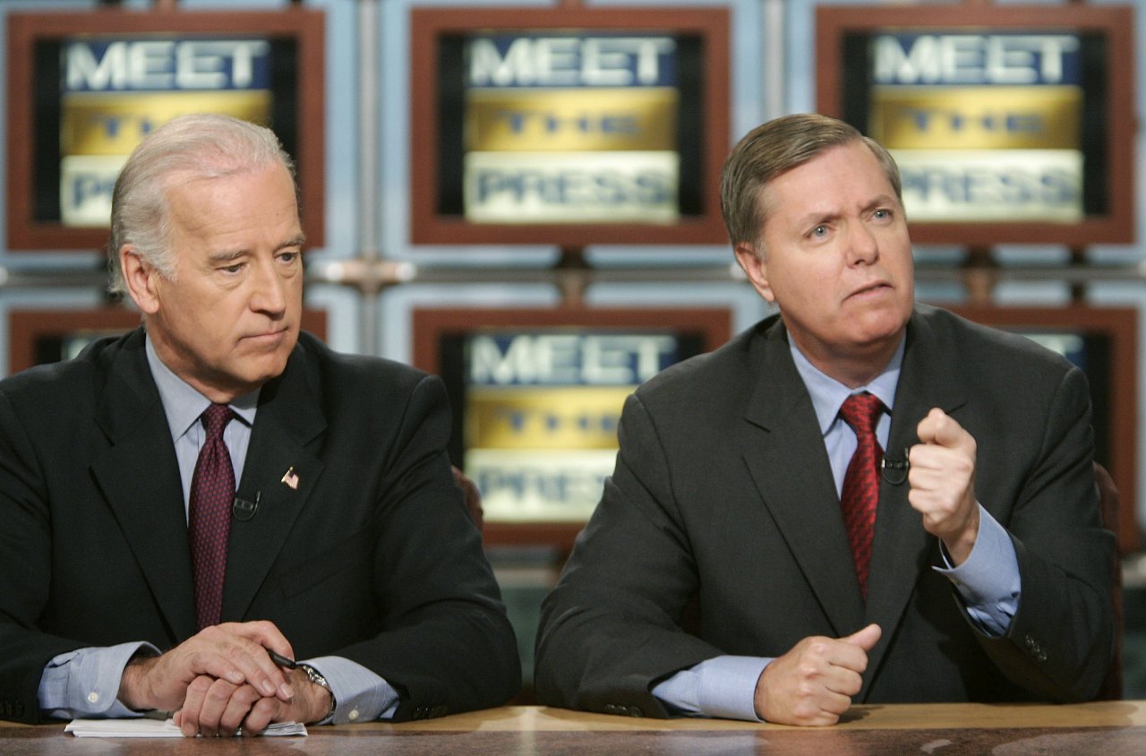 Two men in dark suits sit at a desk in front of multiple 'Meet the Press' monitor displays; the man on the left looks down while the man on the right gestures with his fist.