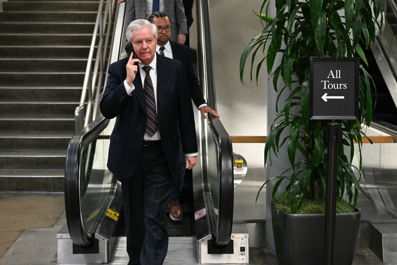 Sen. Lindsey Graham in a navy suit and striped tie talks on a phone while stepping off an escalator on Capitol Hill, with an 'All Tours' sign visible nearby.