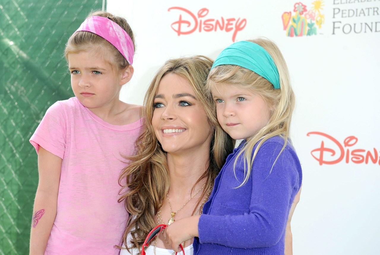 A blonde woman smiling and hugging two young girls — one in pink with a pink headband, one in purple with a teal headband — at a Disney-branded event.