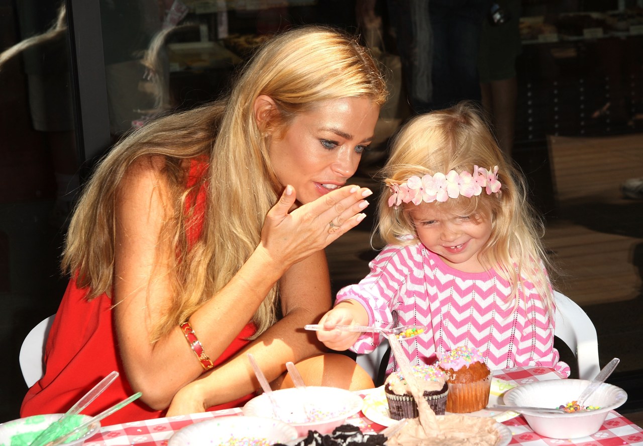 A blonde woman in a red dress watching a smiling blonde toddler in a pink floral headband decorate a cupcake at a picnic table.