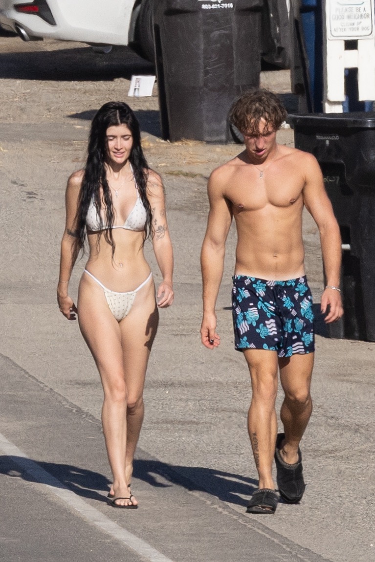 Woman in a white bikini and a shirtless man in blue printed swim trunks walking on a paved path near a beach in Malibu.