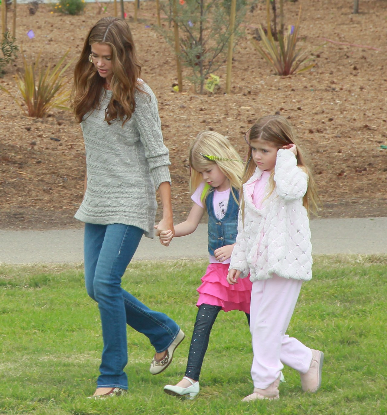 Woman with long brown hair holding hands with a young blonde girl in a pink outfit, walking beside another girl in a white jacket at an outdoor event.